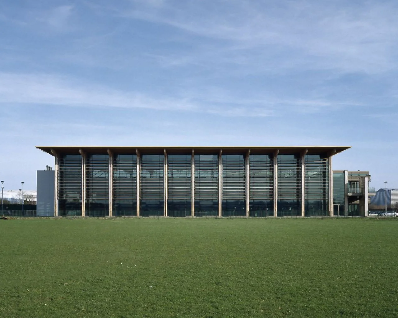 Modern building with glass windows and vertical wooden supports, set against a bright blue sky and green lawn in the foreground.