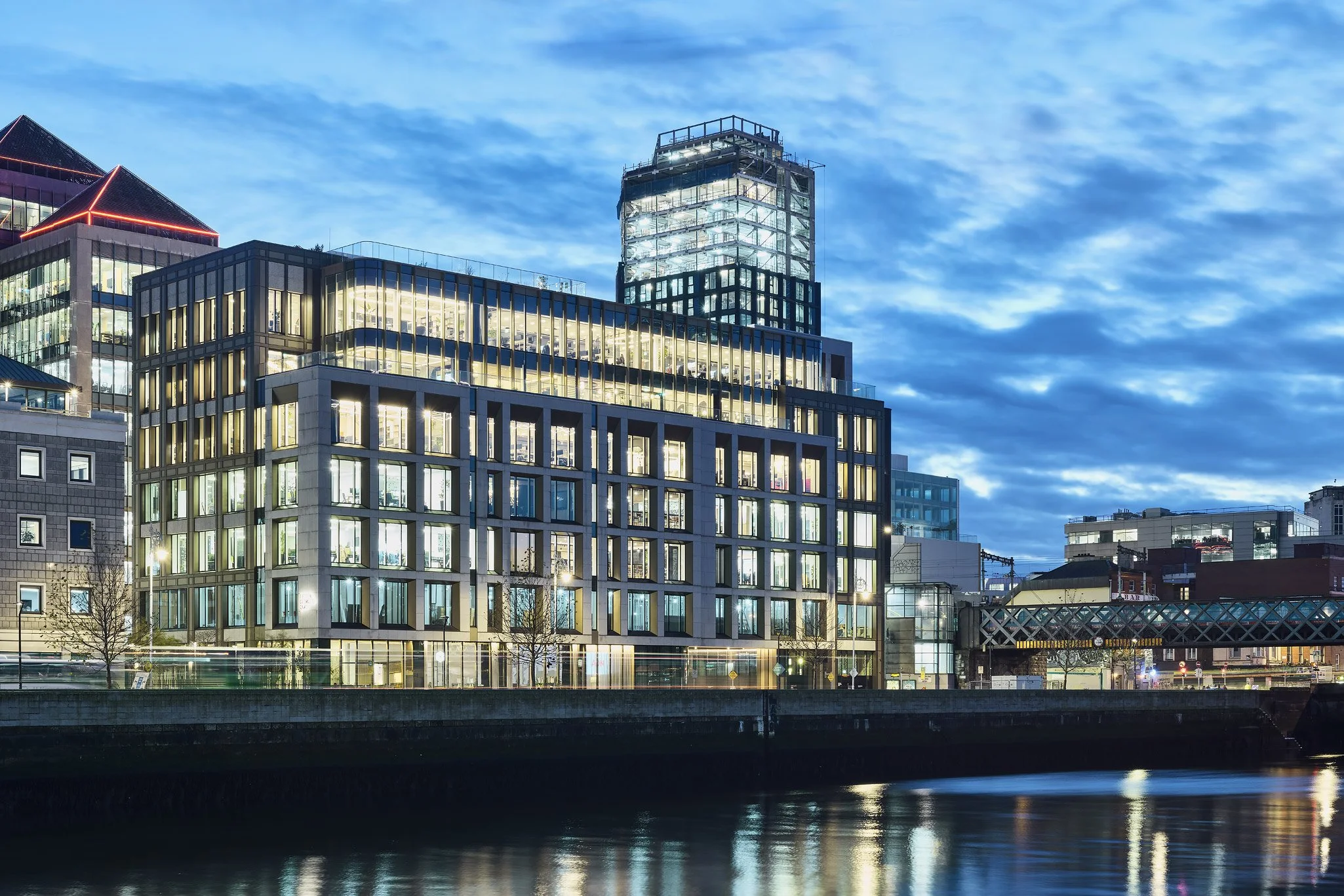 Modern city buildings with illuminated windows reflecting on the water at dusk, with a cloudy sky overhead.