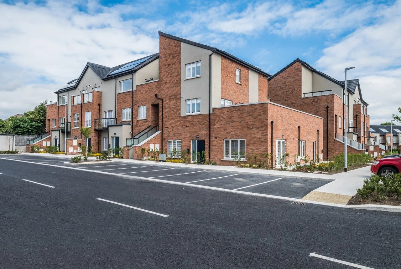 Modern multi-story apartment buildings with brick and beige exteriors, parking lot in the foreground, and a partly cloudy sky.