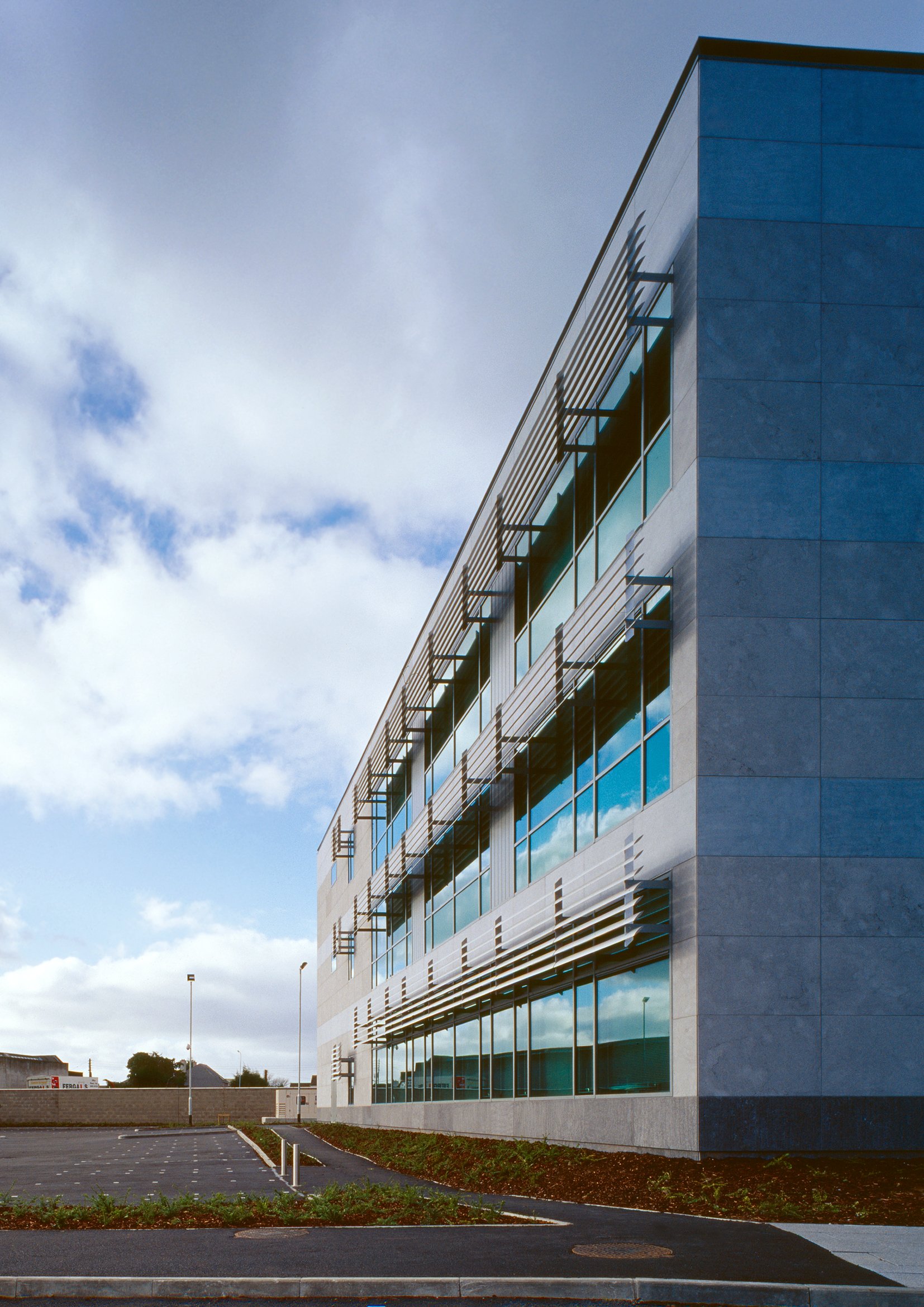 Contemporary office building with large glass windows reflecting the sky, and horizontal metal sunshades on a cloudy day.