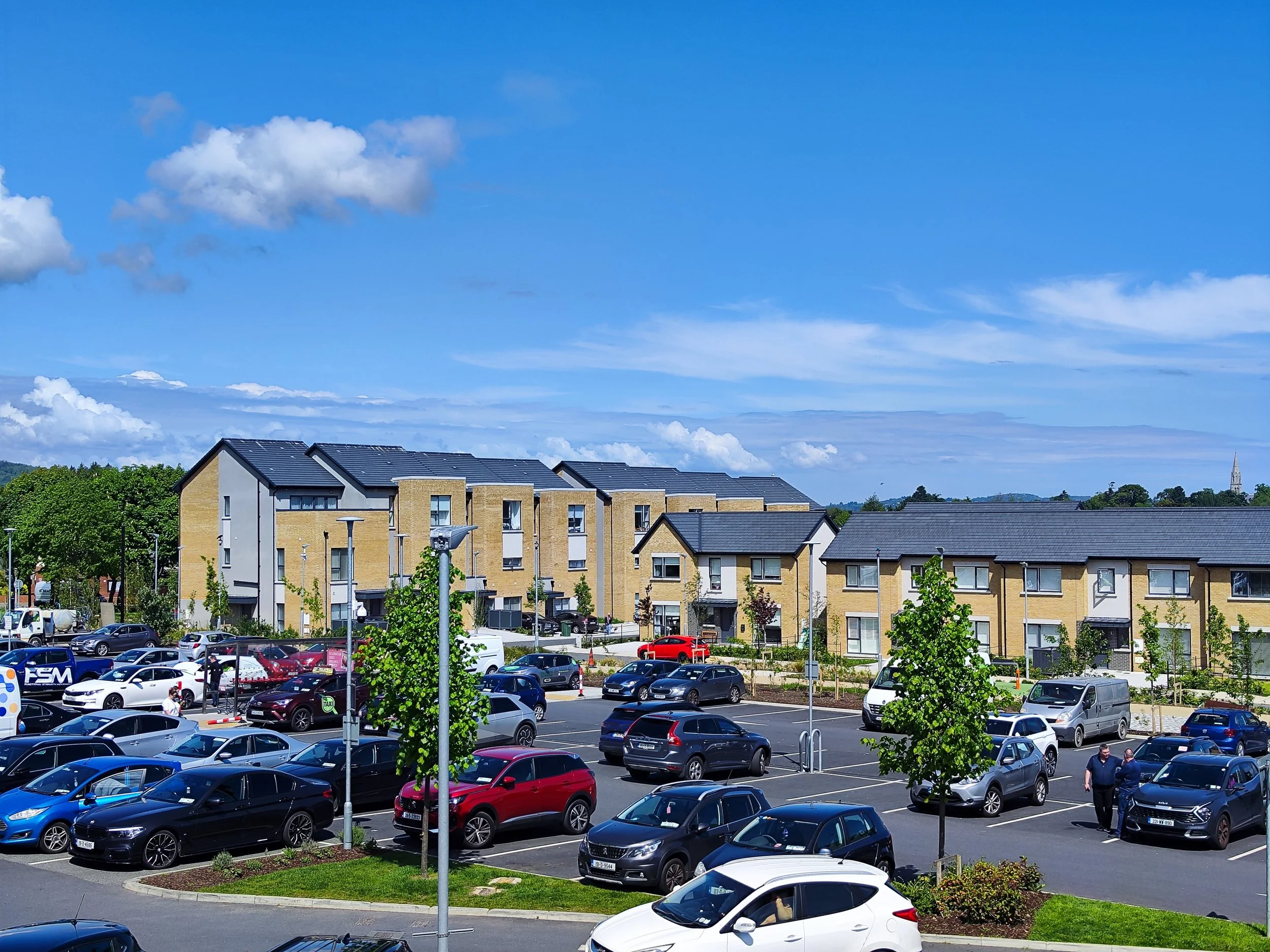 A parking lot with multiple cars, surrounded by trees and modern residential buildings under a blue sky with some clouds.