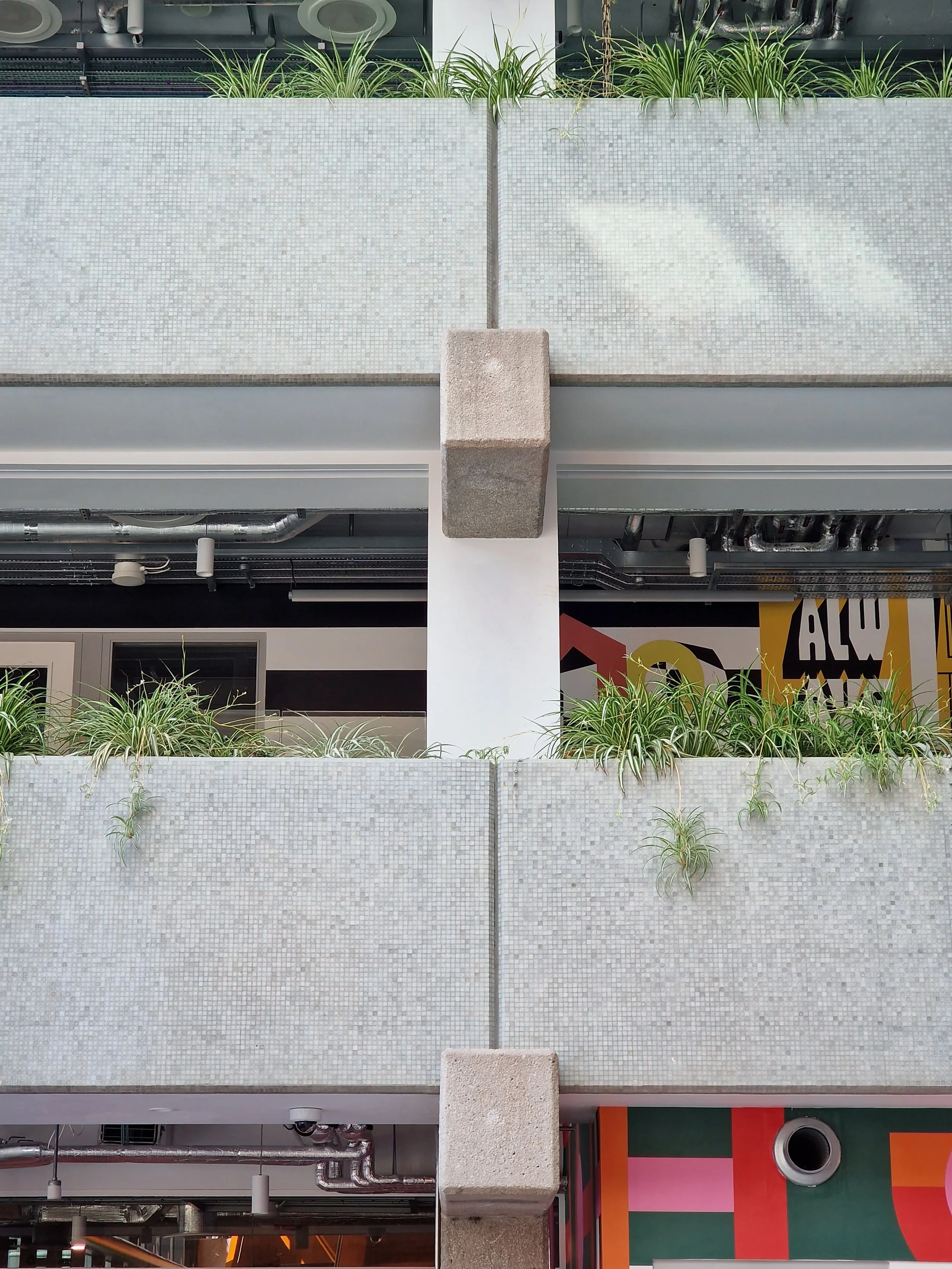 Two levels of outdoor space with tiled concrete barriers and green plants on top, featuring a central white column and concrete support blocks.