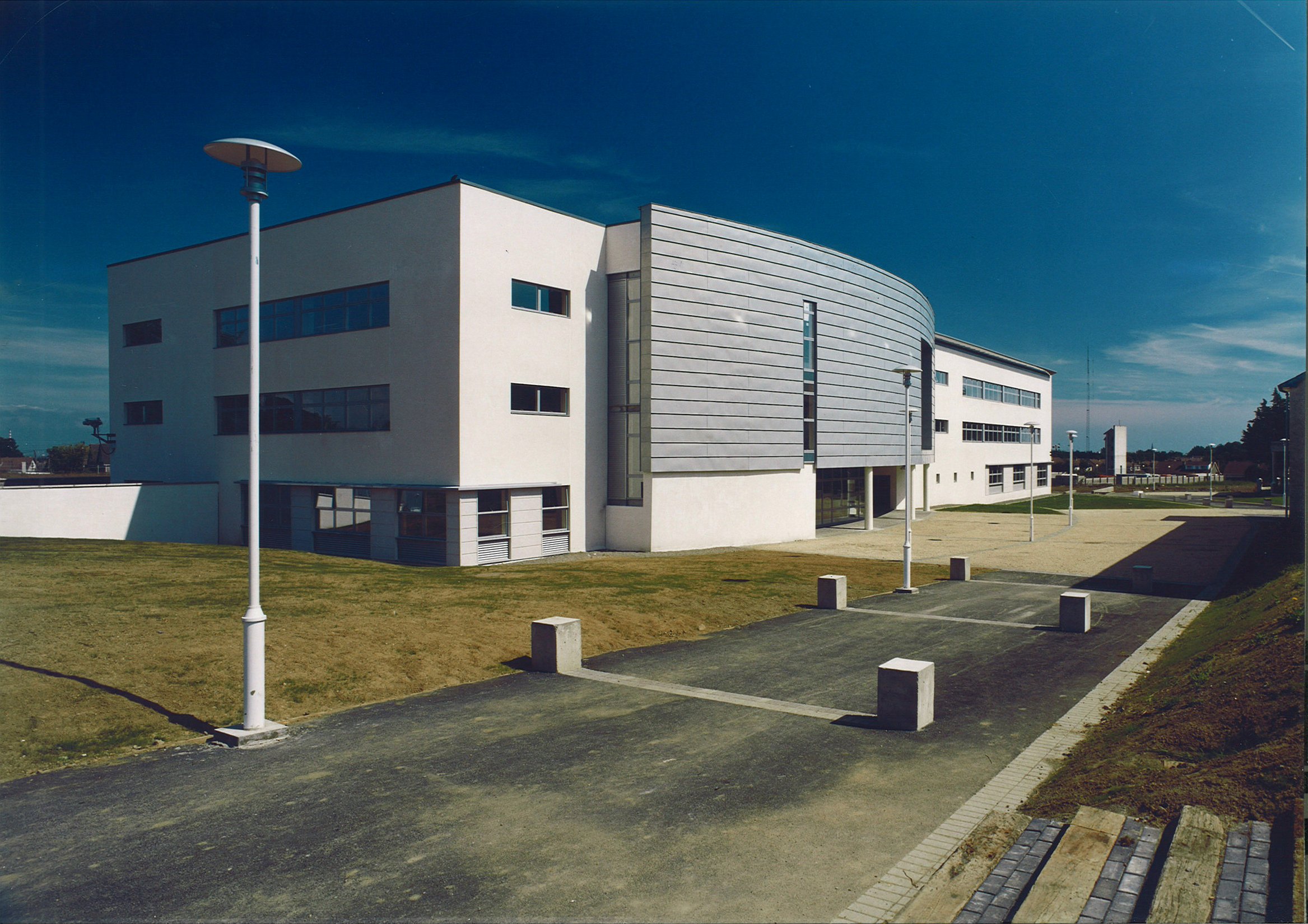 Modern white building with curved gray facade, windows, and a well-maintained grassy area with lamp posts and parking blocks.
