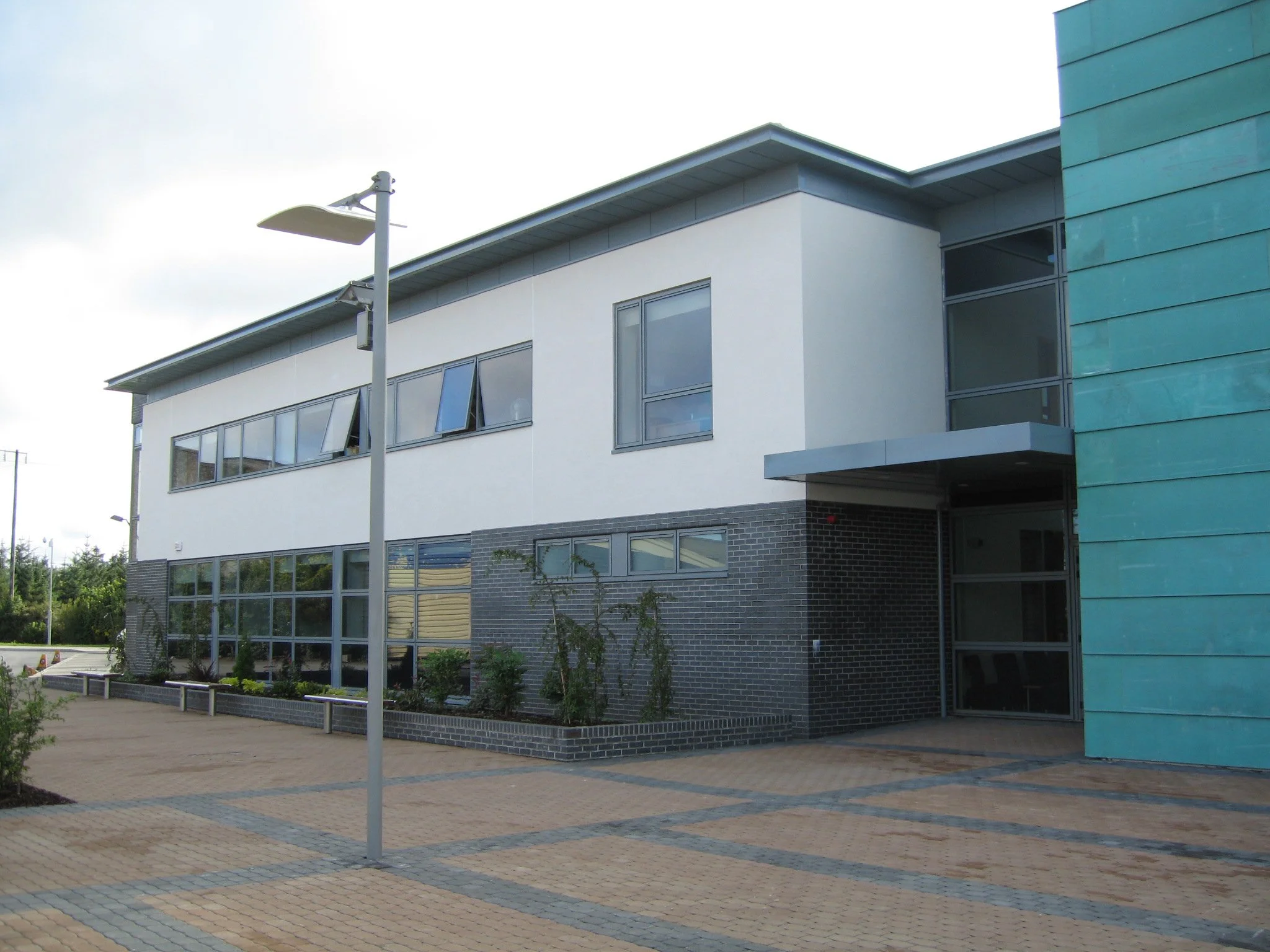 Modern building with large glass windows, gray brick and white walls, and a green side panel, with a paved walkway and a streetlamp in the foreground.
