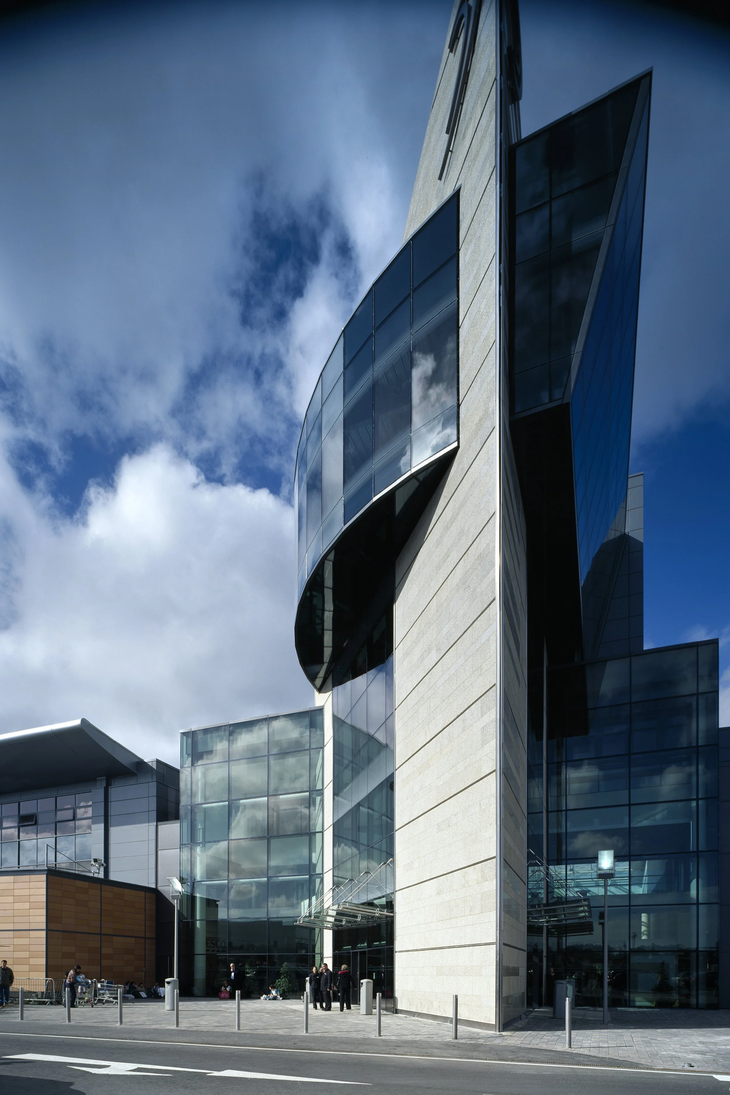 Modern glass and concrete building with people standing outside under a partly cloudy sky.