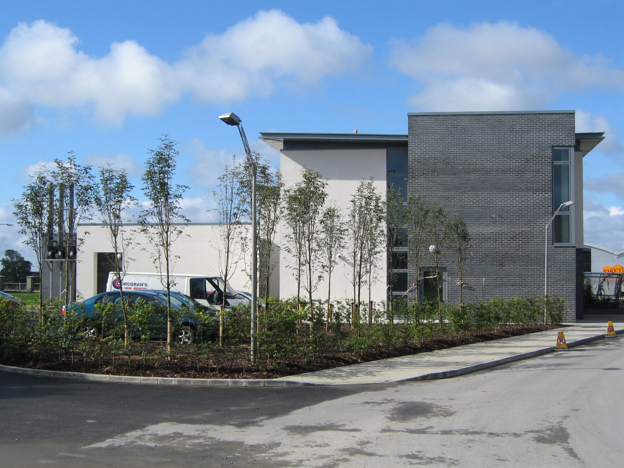 Modern building with white and gray brick exterior, surrounded by trees and a parking lot with a blue car and a white van.