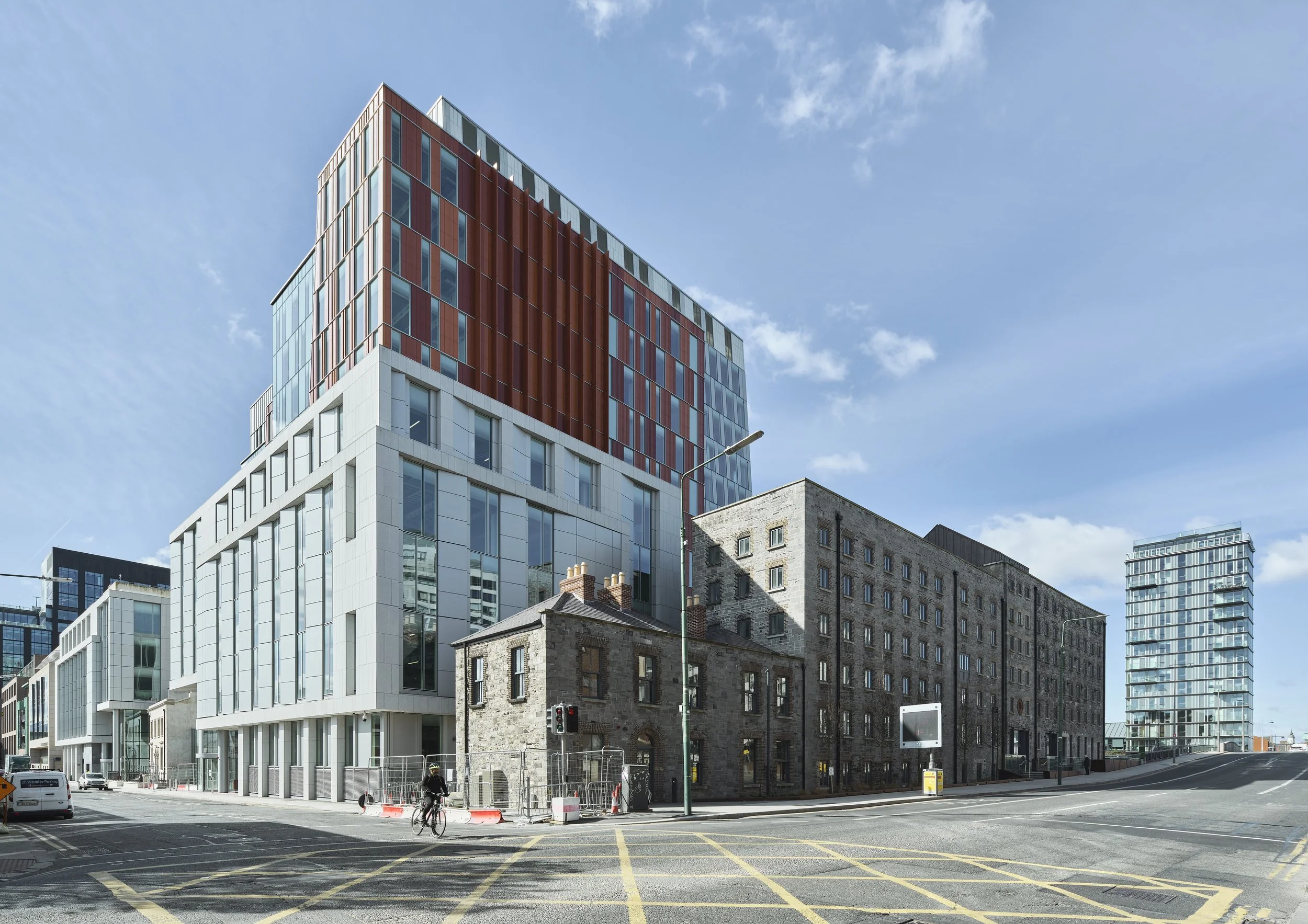 A city street corner featuring a modern office building with glass windows and red panels, adjacent to a historic stone building. In the background, more contemporary buildings can be seen. A cyclist rides on the road, and traffic signs are visible.