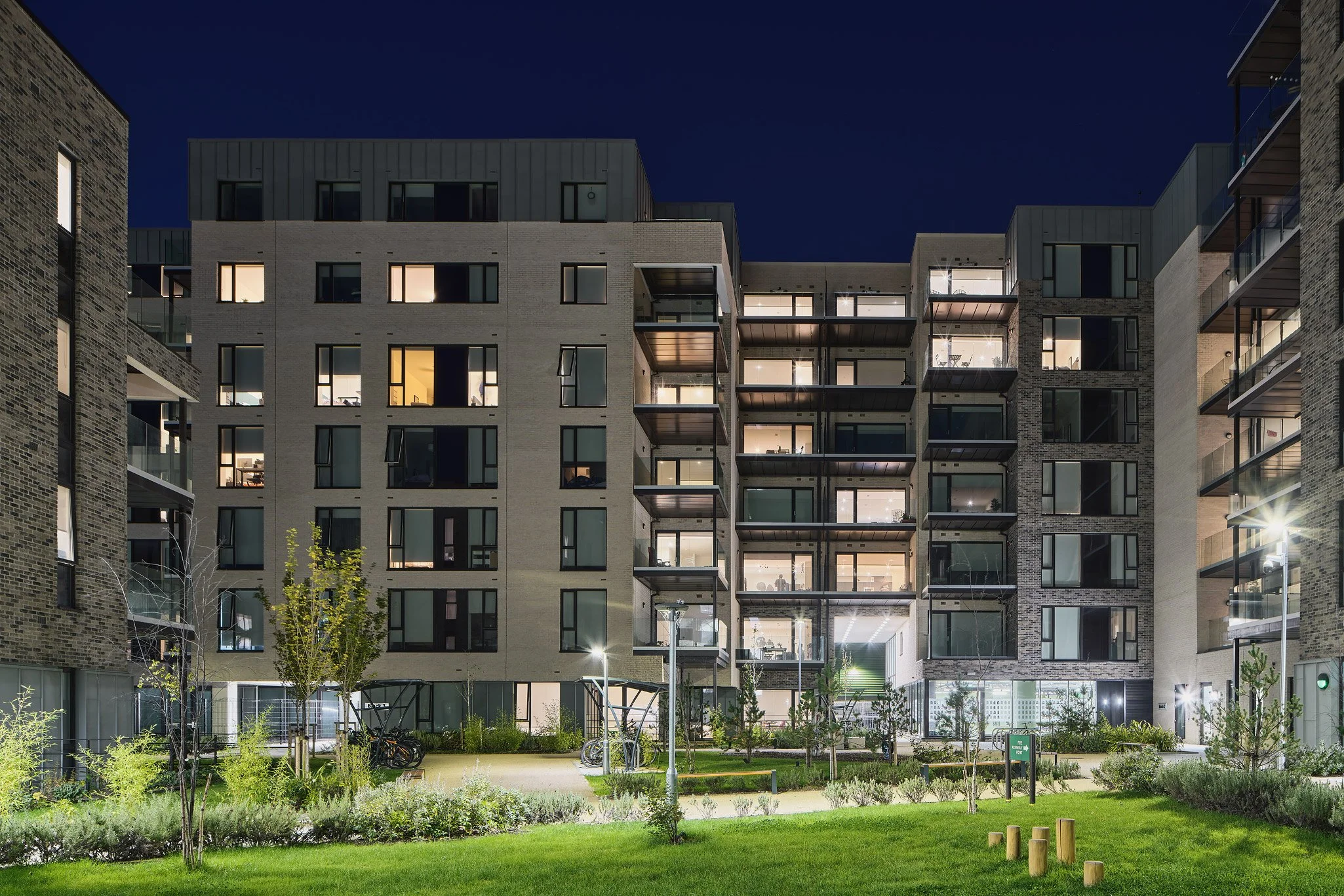 Night view of a modern apartment complex with lit windows and outdoor lighting, surrounded by a landscaped yard with trees, grass, and a pathway.