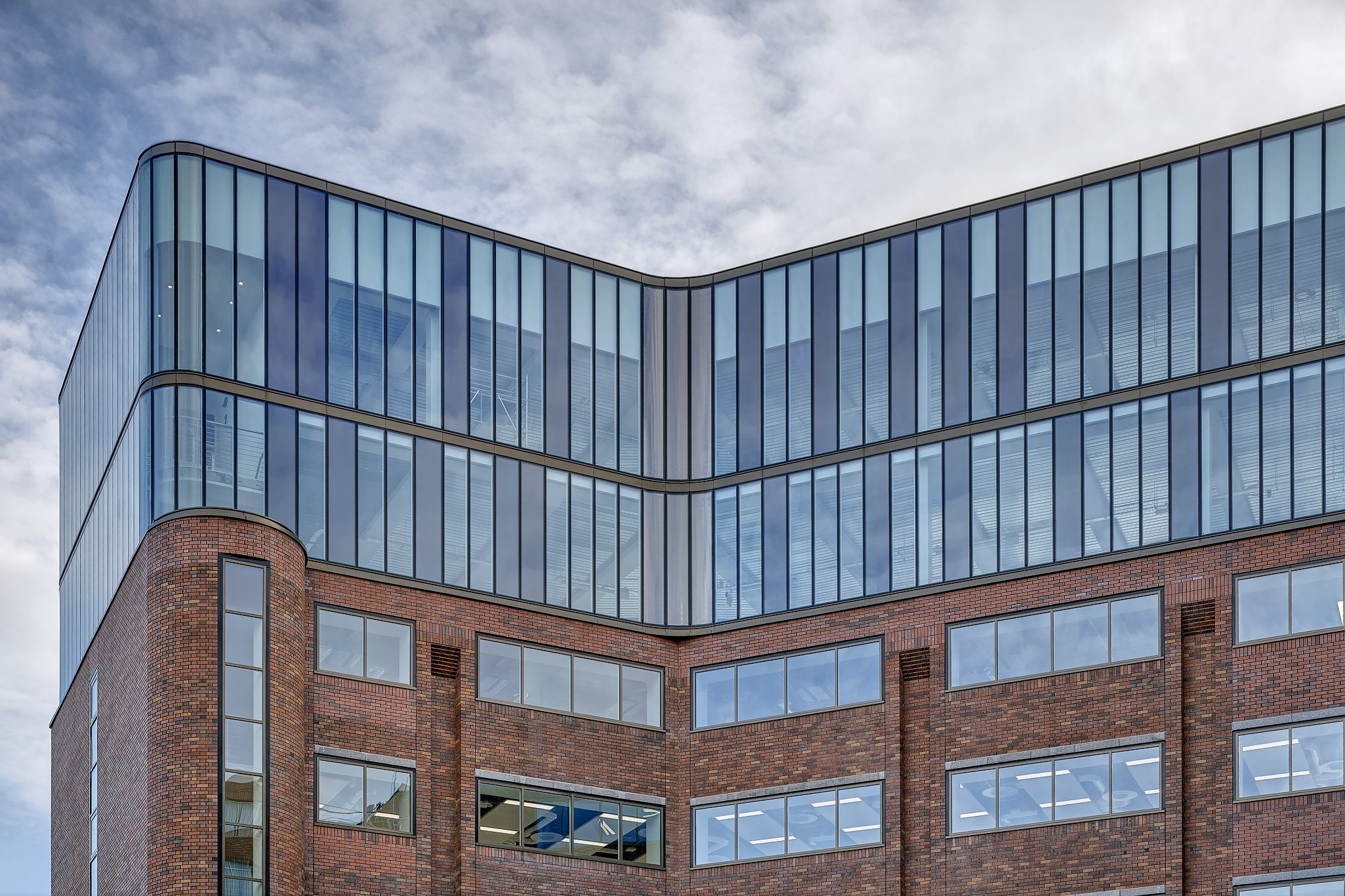 A modern building with a lower section of brick and glass windows, and an upper section of glass panels reflecting the sky.