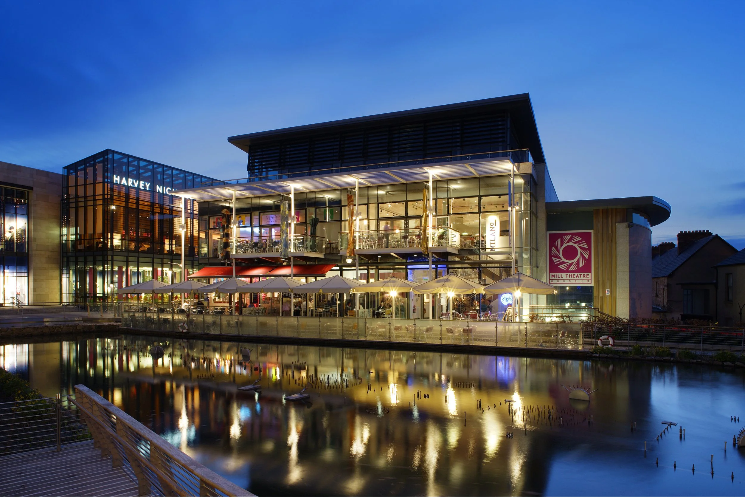 A modern multi-story building with glass windows and a restaurant on the ground floor, illuminated at dusk. There are outdoor umbrellas and seating along a water feature reflecting the building's lights.