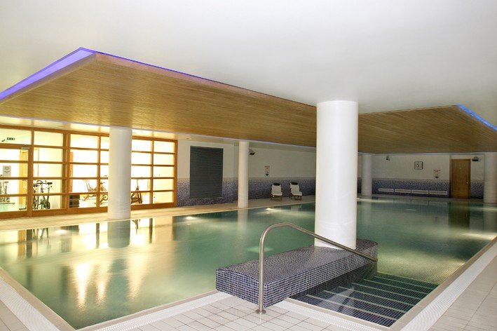 Indoor swimming pool area with white columns, wooden ceiling, tiled flooring, lounge chairs, and a glass wall allowing natural light.