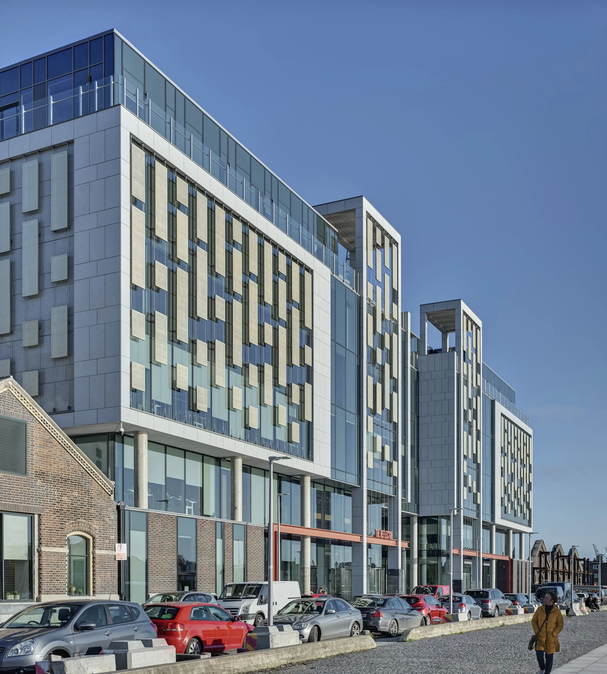 Modern multi-story office building with glass windows and decorative panels, parked cars in front, and a person walking nearby under a clear blue sky.
