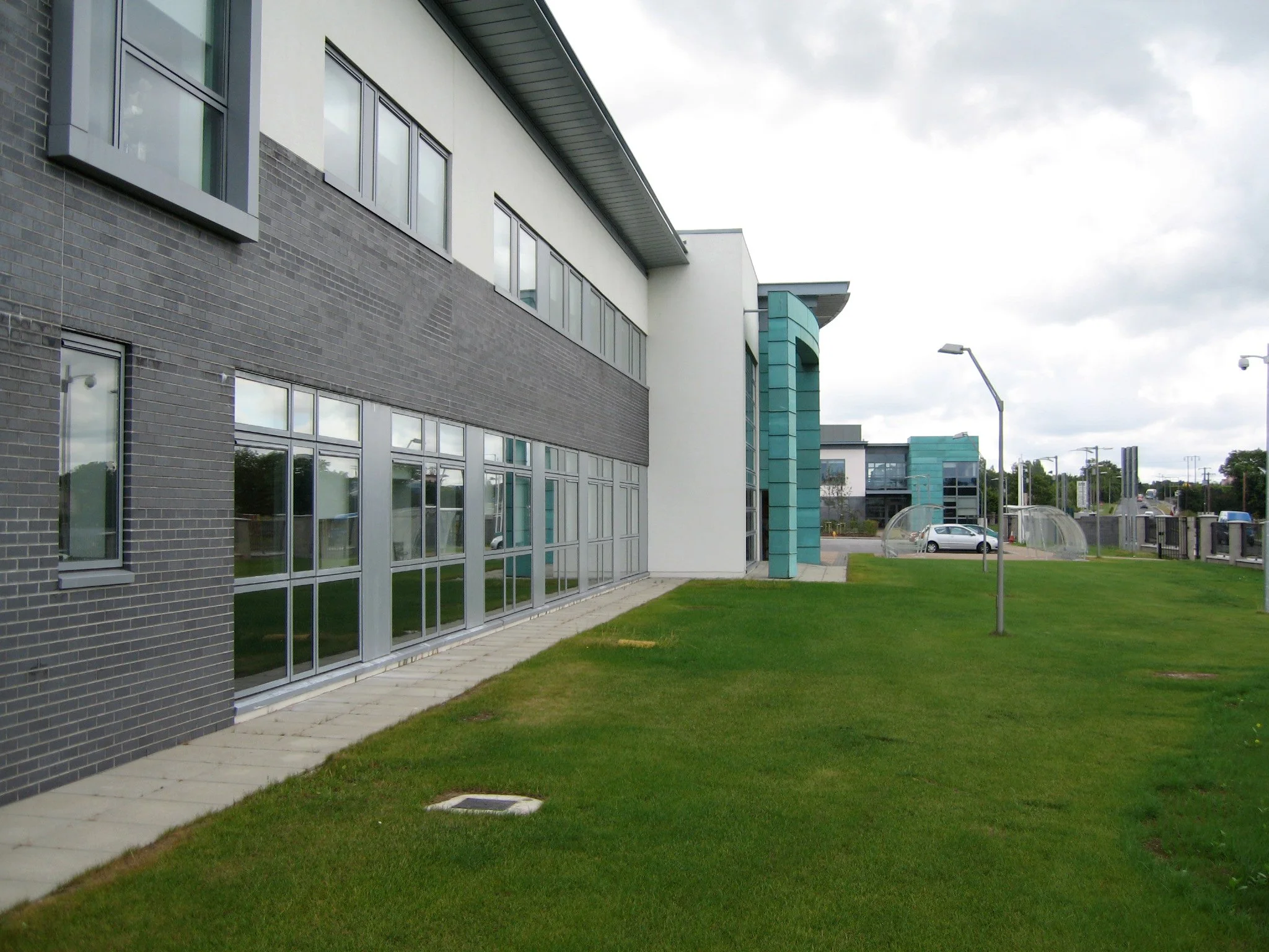 modern office building with large glass windows and a green lawn outside, cloudy sky overhead, street lights and cars in the background.