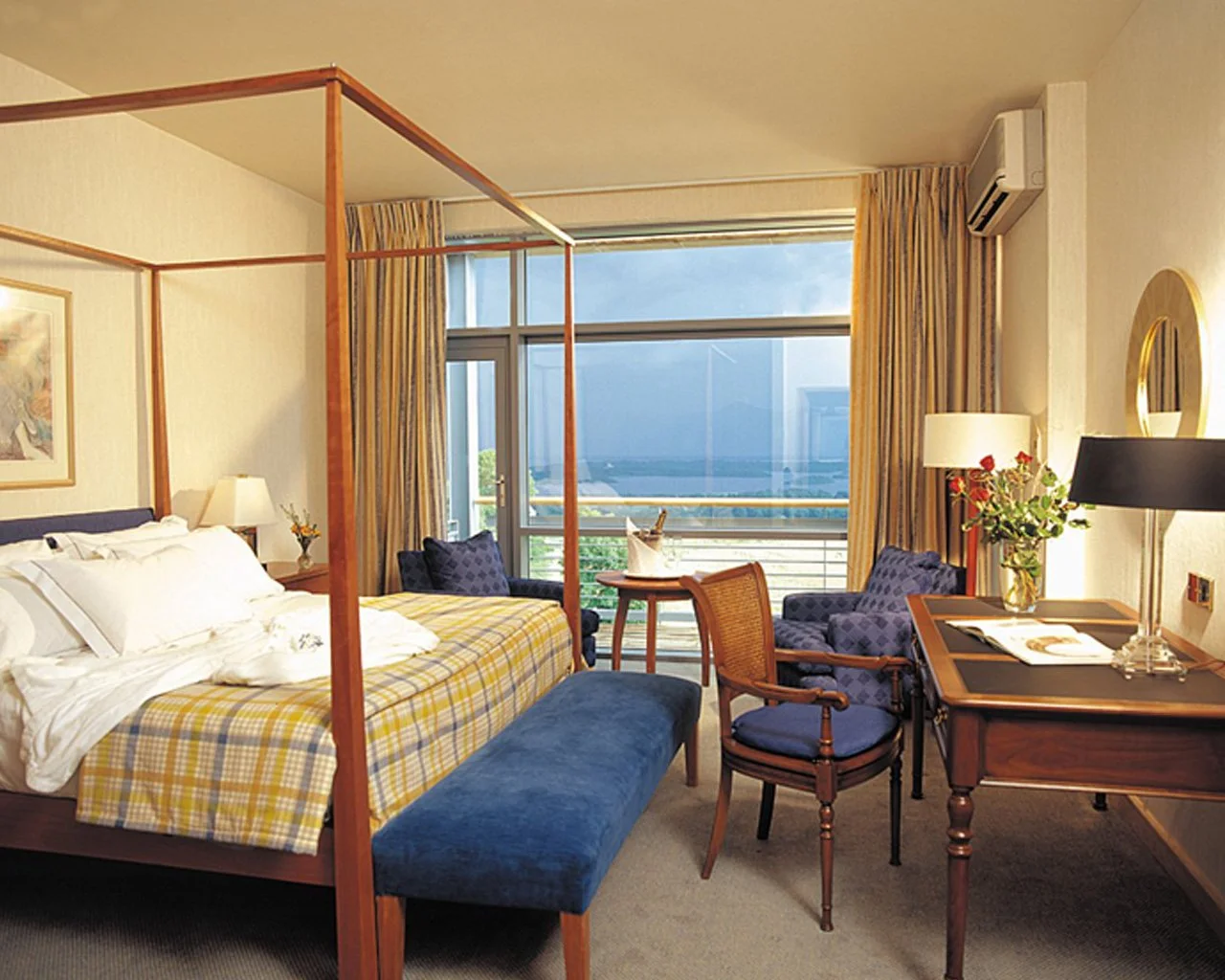Hotel room with a four-poster bed, blue upholstered bench, side table with lamp, two blue armchairs, and a wooden desk with a black lamp and a vase of flowers. Large window with yellow curtains shows cityscape view.