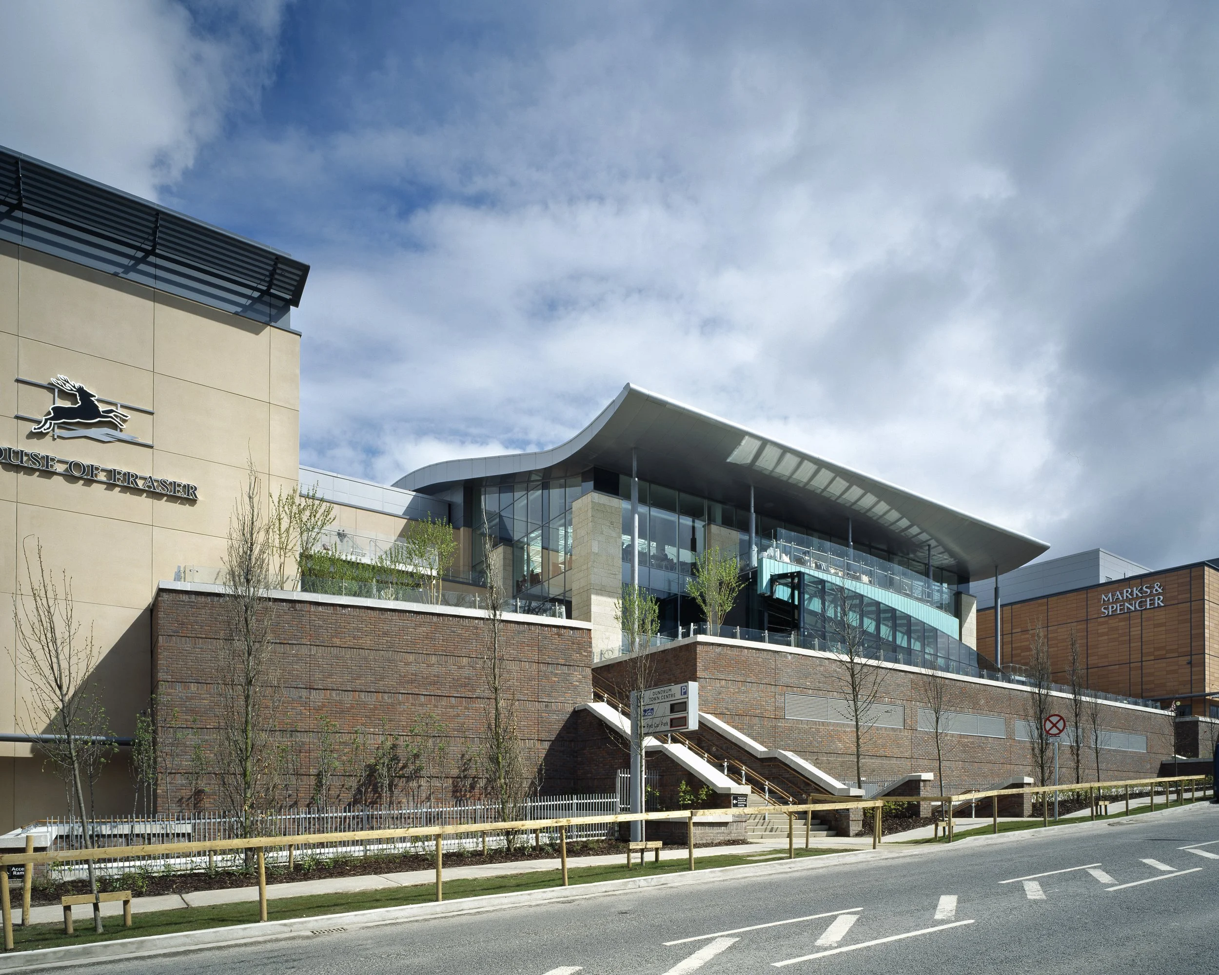 Modern shopping mall with a glass facade, brick walls, and surrounding trees, featuring signs for House of Fraser and Marks & Spencer.