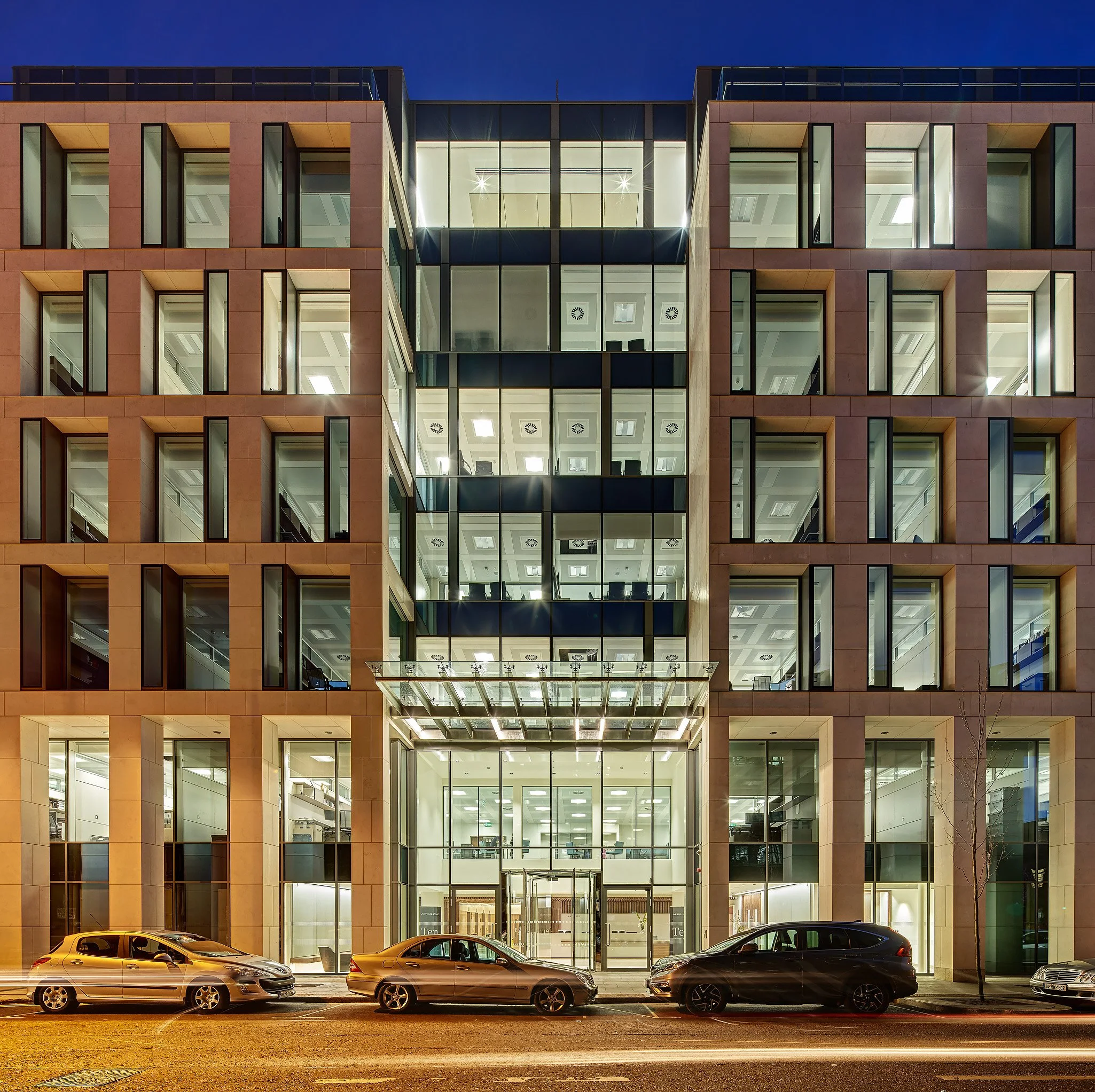 Modern multi-story office building with glass windows, illuminated interior, and cars parked outside at night.