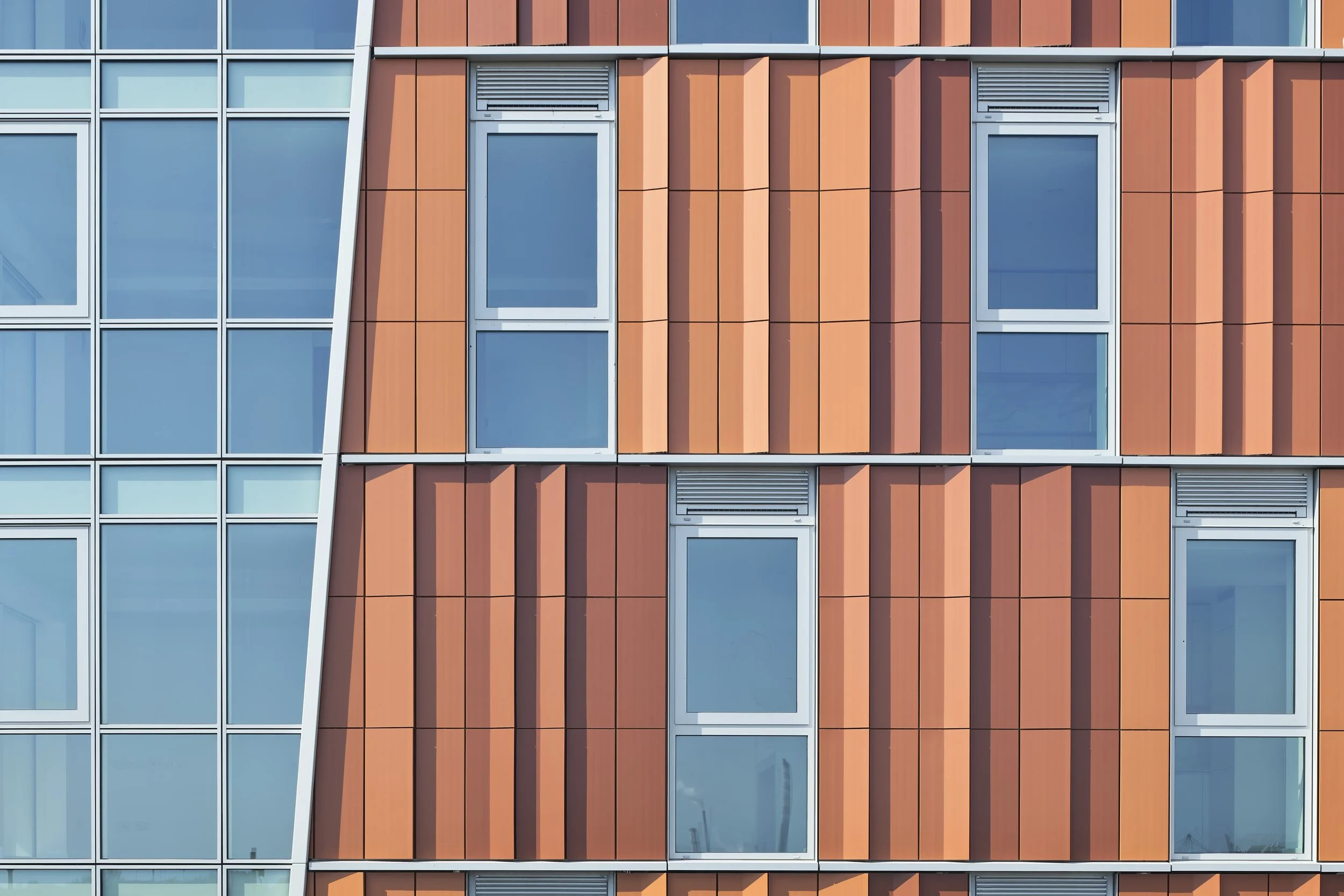 Close-up view of a modern building's facade with orange panels and glass windows arranged in a grid pattern, with some windows having vents.