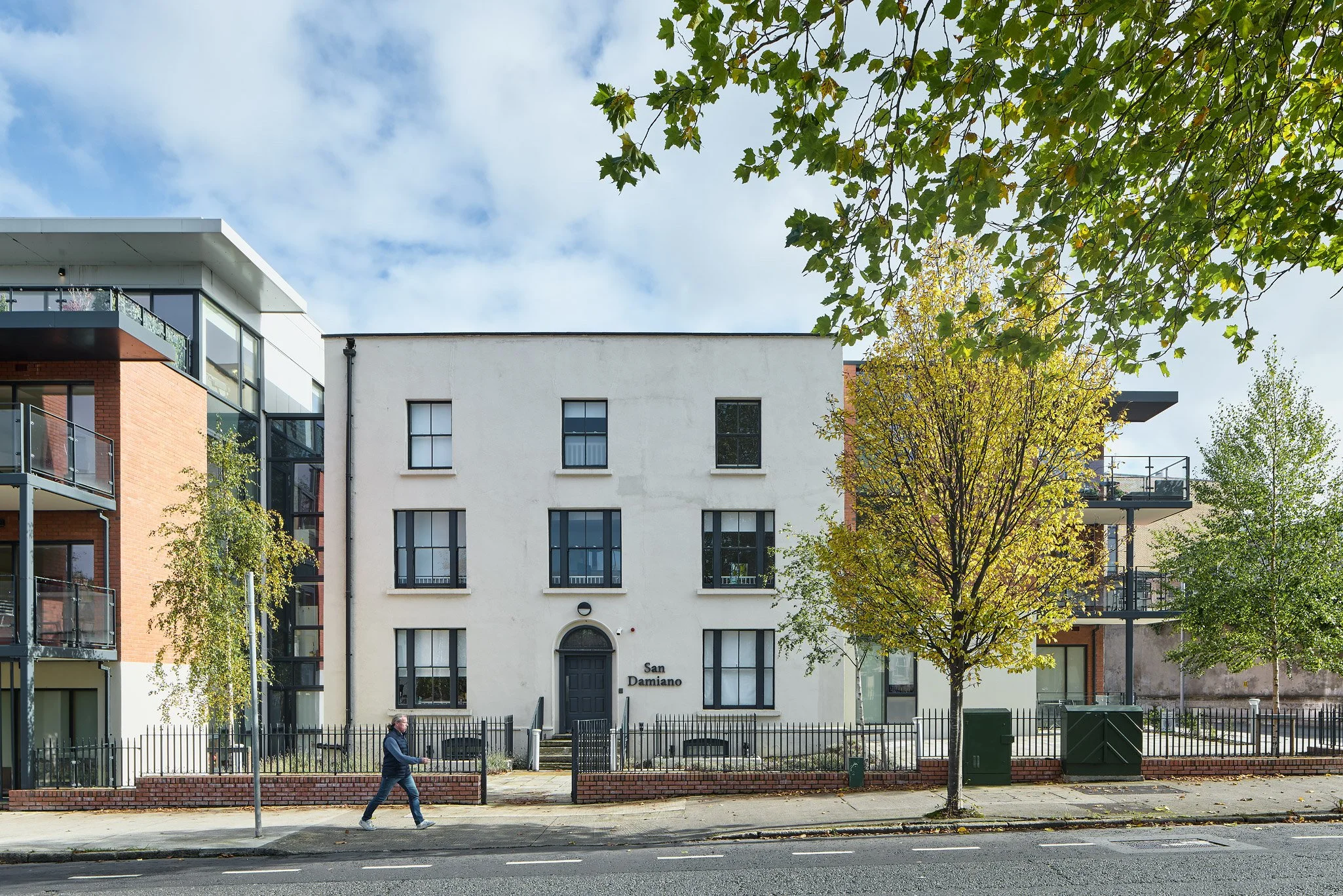 A white three-story building with black window frames and a black door, labeled 'San Damiano', is flanked by modern residential buildings on both sides. There are trees with fall foliage in front, and a man is walking on the sidewalk.