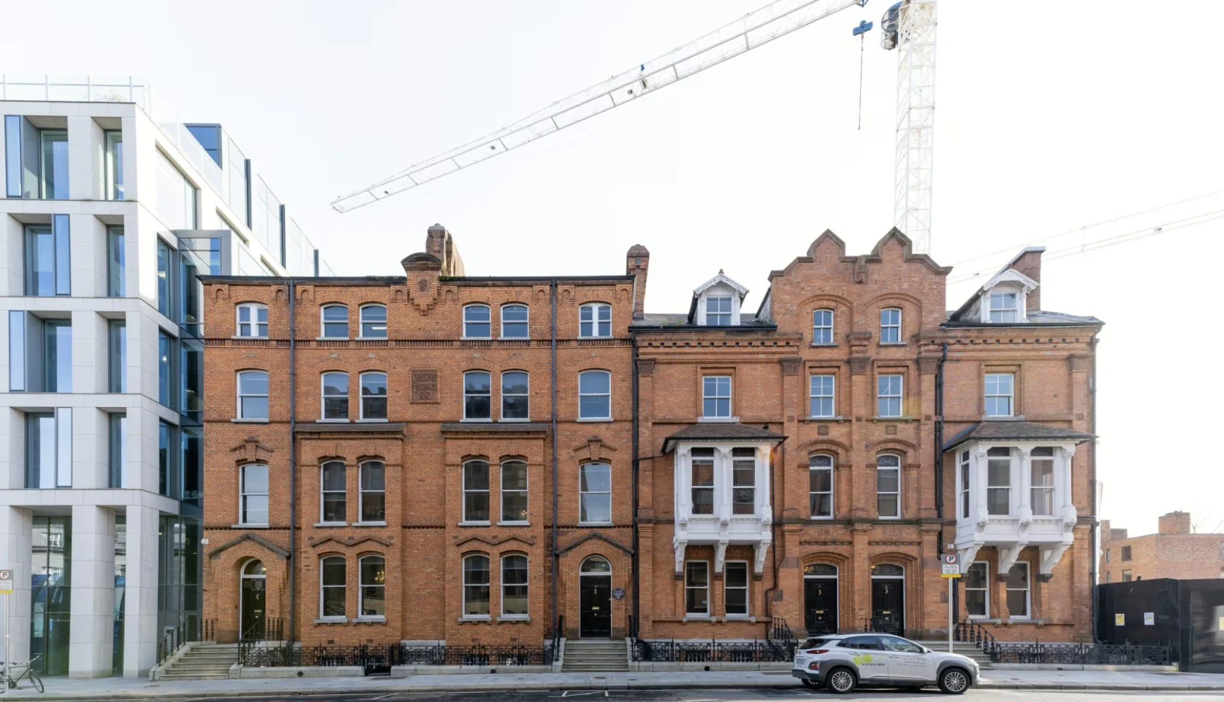 A row of historic red brick buildings with ornate architectural details next to a modern white and glass building, with a construction crane in the background and a parked gray vehicle in front.