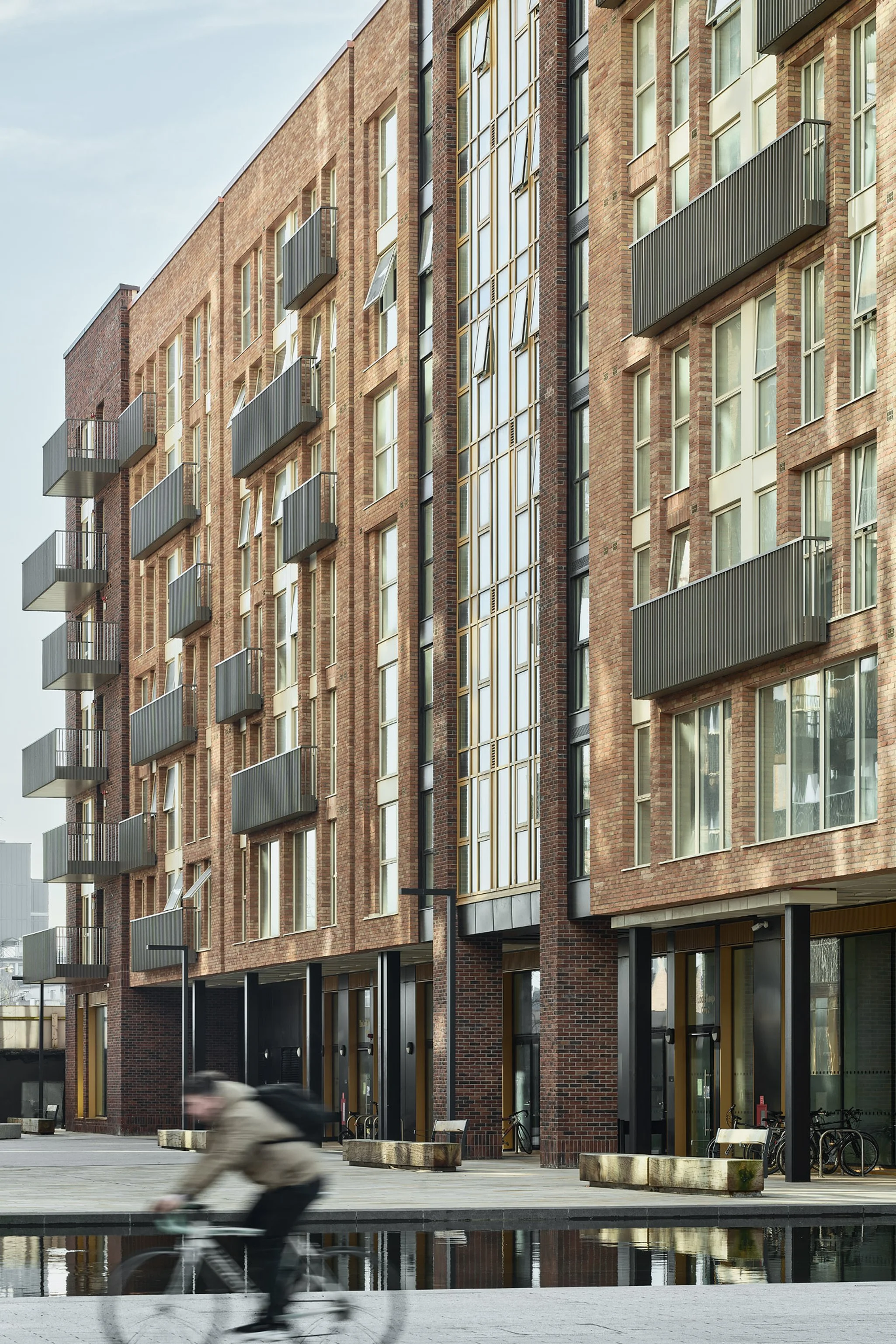 A modern brick apartment building with multiple floors, balconies, and large glass windows. A person riding a bicycle is blurred in motion in the foreground, with benches and bicycles parked near the building. The scene is outdoors with a paved area and a small water feature or reflective surface.