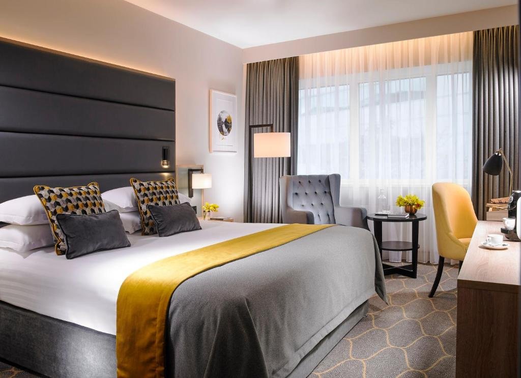 A modern hotel room with a large bed, dark gray upholstered headboard, and decorative cushions. There is a sitting area with a gray armchair, a yellow chair, and a small round table with flowers. A desk with a yellow chair, coffee maker, and cups is near the window with sheer and dark gray curtains.