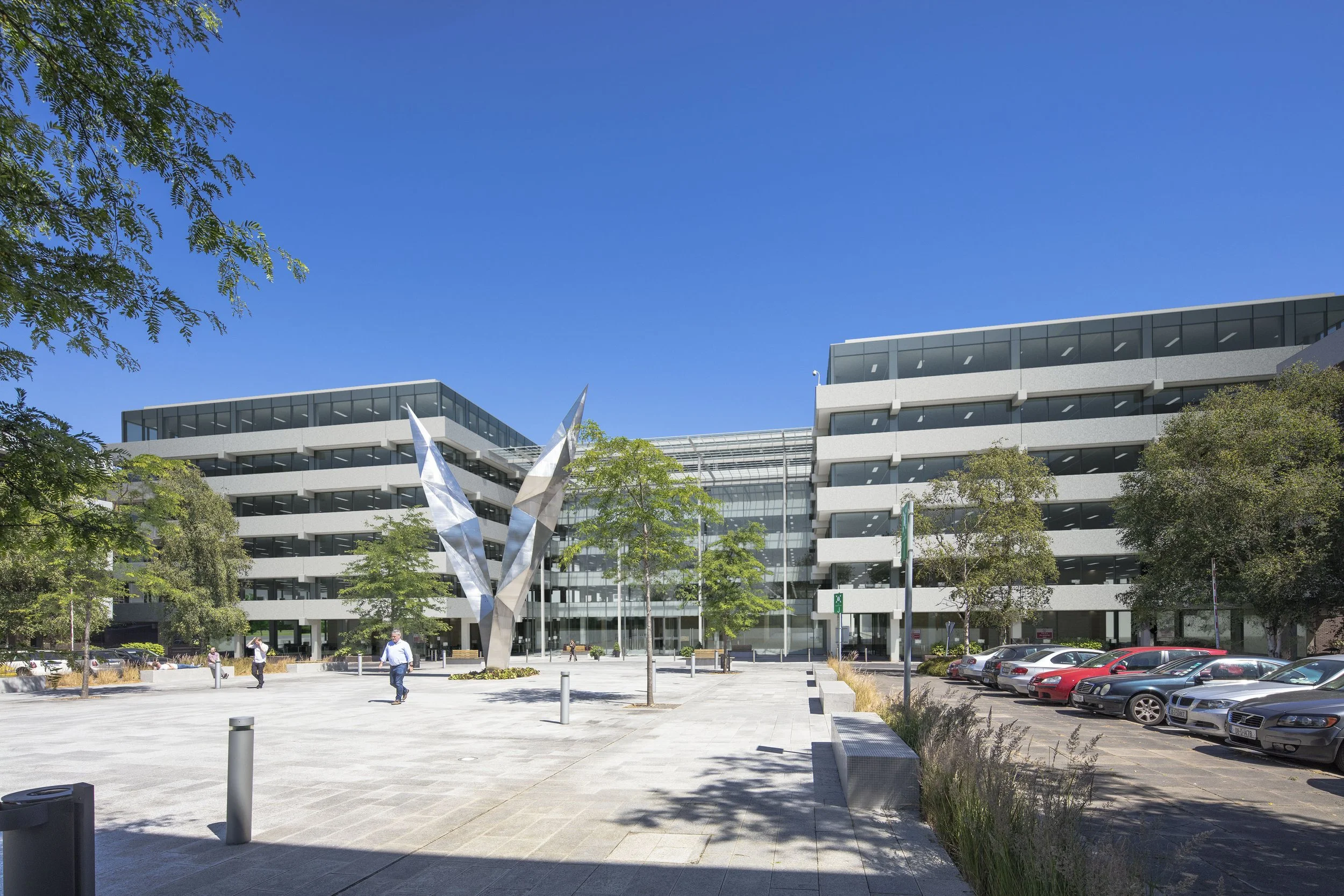 Modern office building with glass and concrete facade, a parking lot with cars, a large metallic sculpture in the foreground, and a clear blue sky overhead.