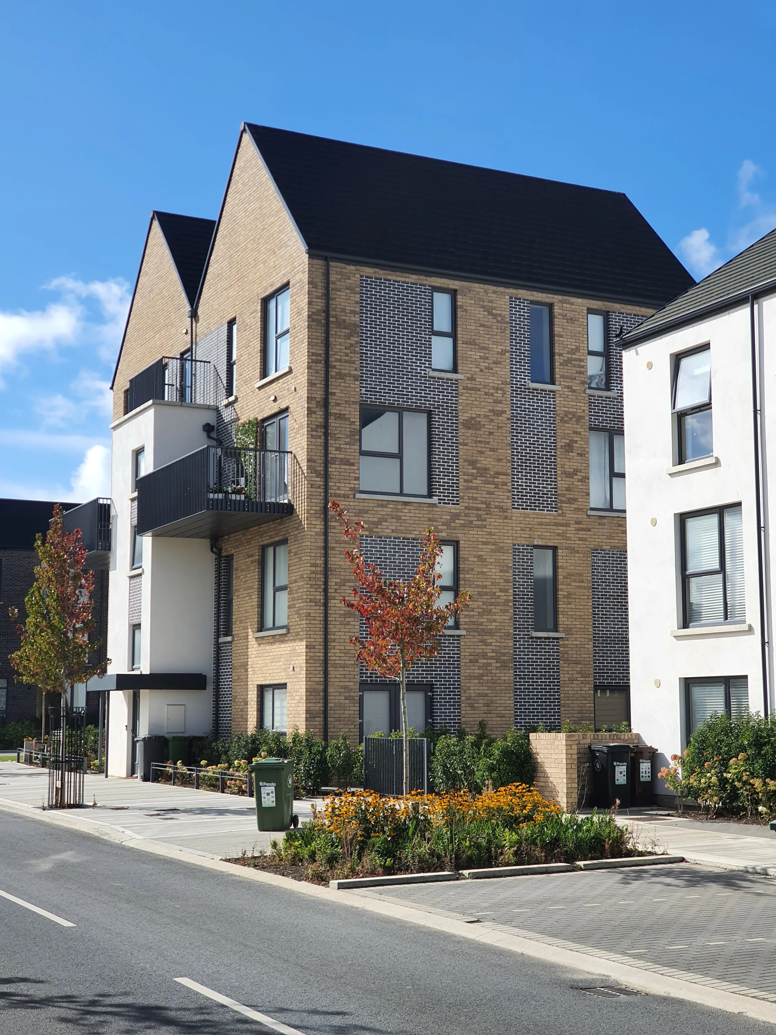 Modern multi-story residential building with balconies, trees, and a landscaped front yard, under a partly cloudy blue sky.