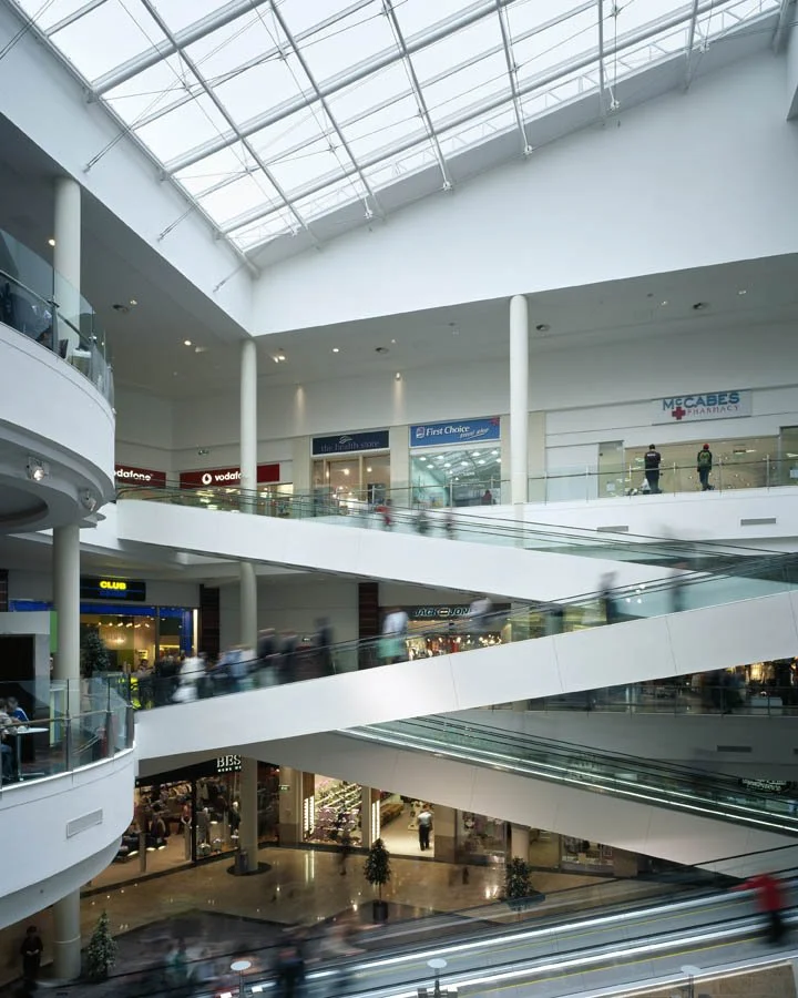 Interior of a multi-level shopping mall with escalators, storefronts, and people walking, including a pharmacy on the upper level under a glass ceiling.