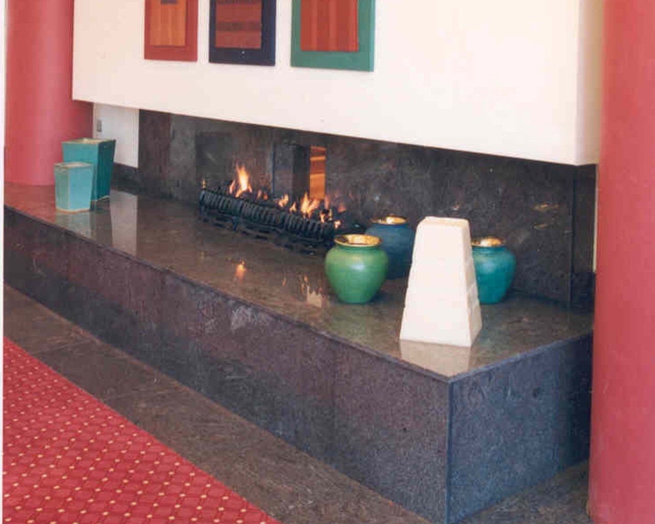 Indoor fireplace with fire burning, surrounded by ceramic vases and decorative objects, in a room with pink walls and red patterned carpet.