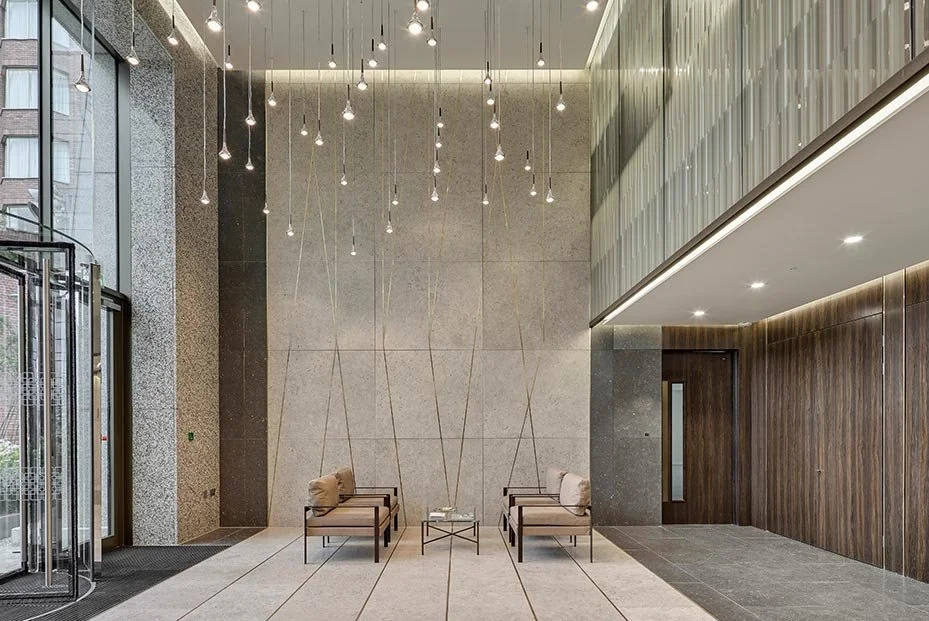 Modern hotel lobby with high ceiling, hanging light fixtures, large glass windows, and minimalist seating area with two chairs and a small table.
