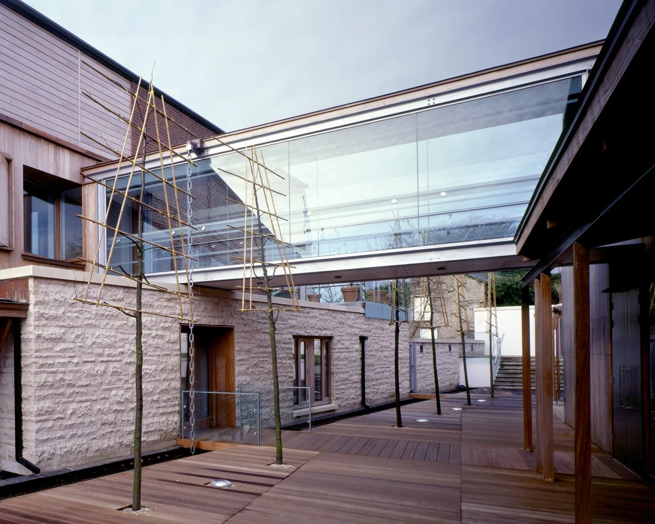 Modern residential courtyard with wooden decking, young trees supported by bamboo scaffolding, and a glass walkway connecting two buildings.