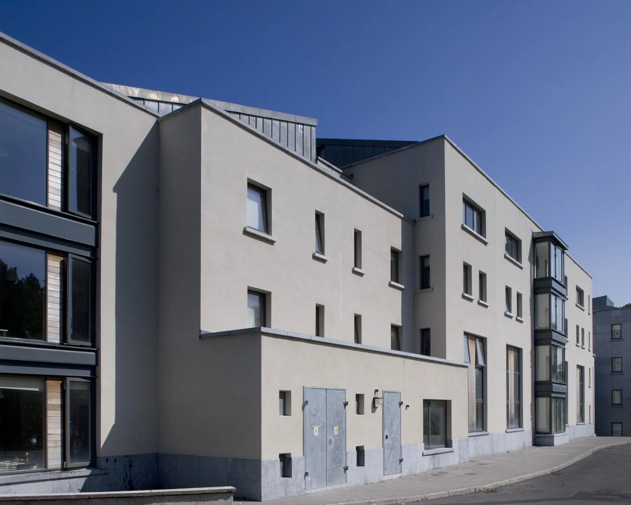 A modern beige apartment building with multiple windows and a small sidewalk in front, under a clear blue sky.