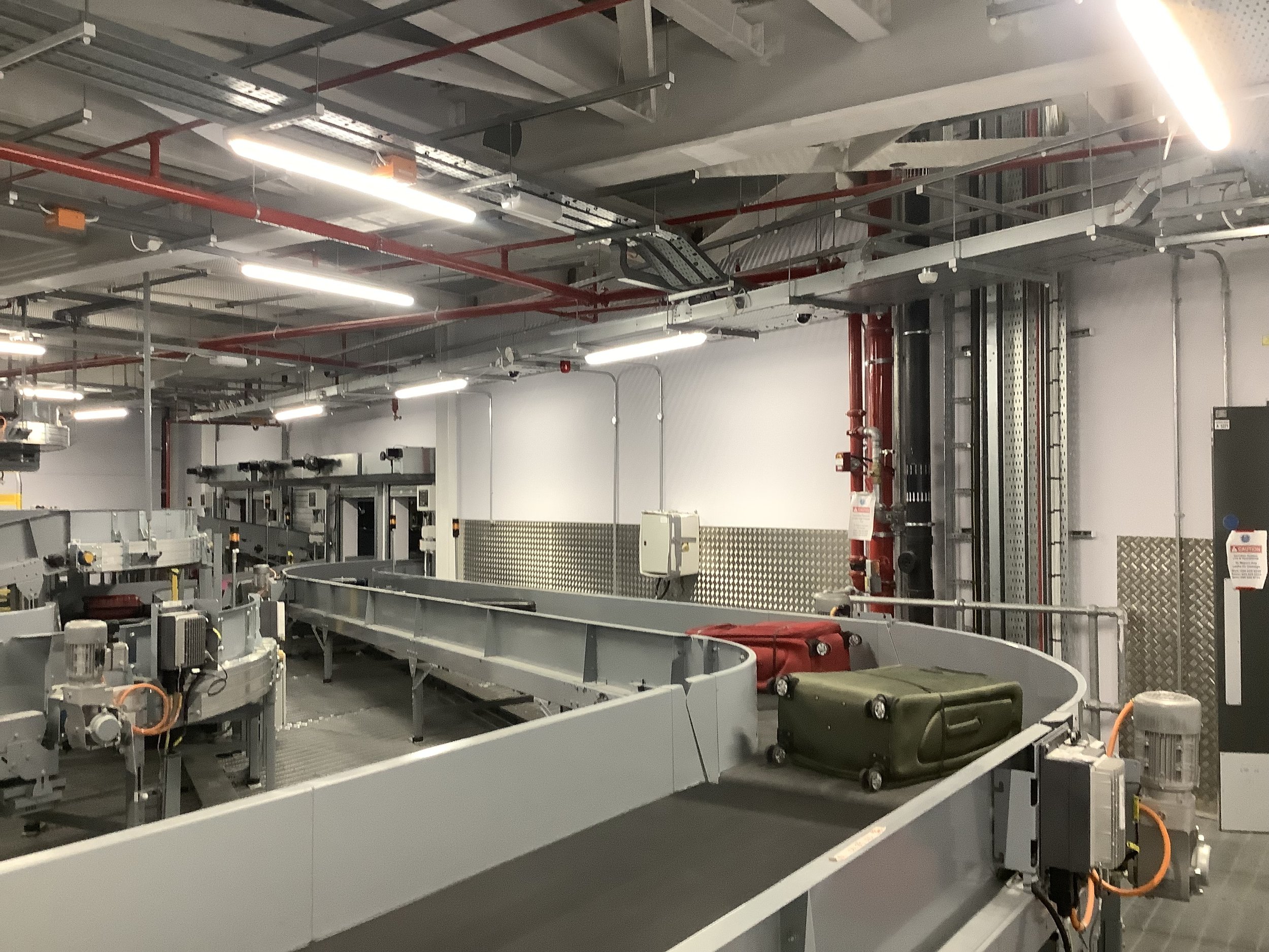 Empty airport baggage claim conveyor belt with two pieces of luggage, one green and one red, and industrial ceiling with lighting and pipes.