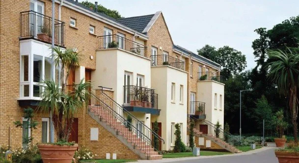 A multi-story residential building with brick and painted exterior, featuring balconies, stairs, and surrounding greenery.