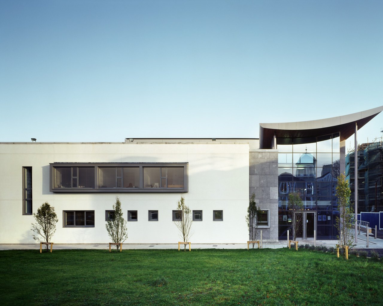 Modern building with a white facade, large windows, and a curved roof section, surrounded by a green lawn and young trees.
