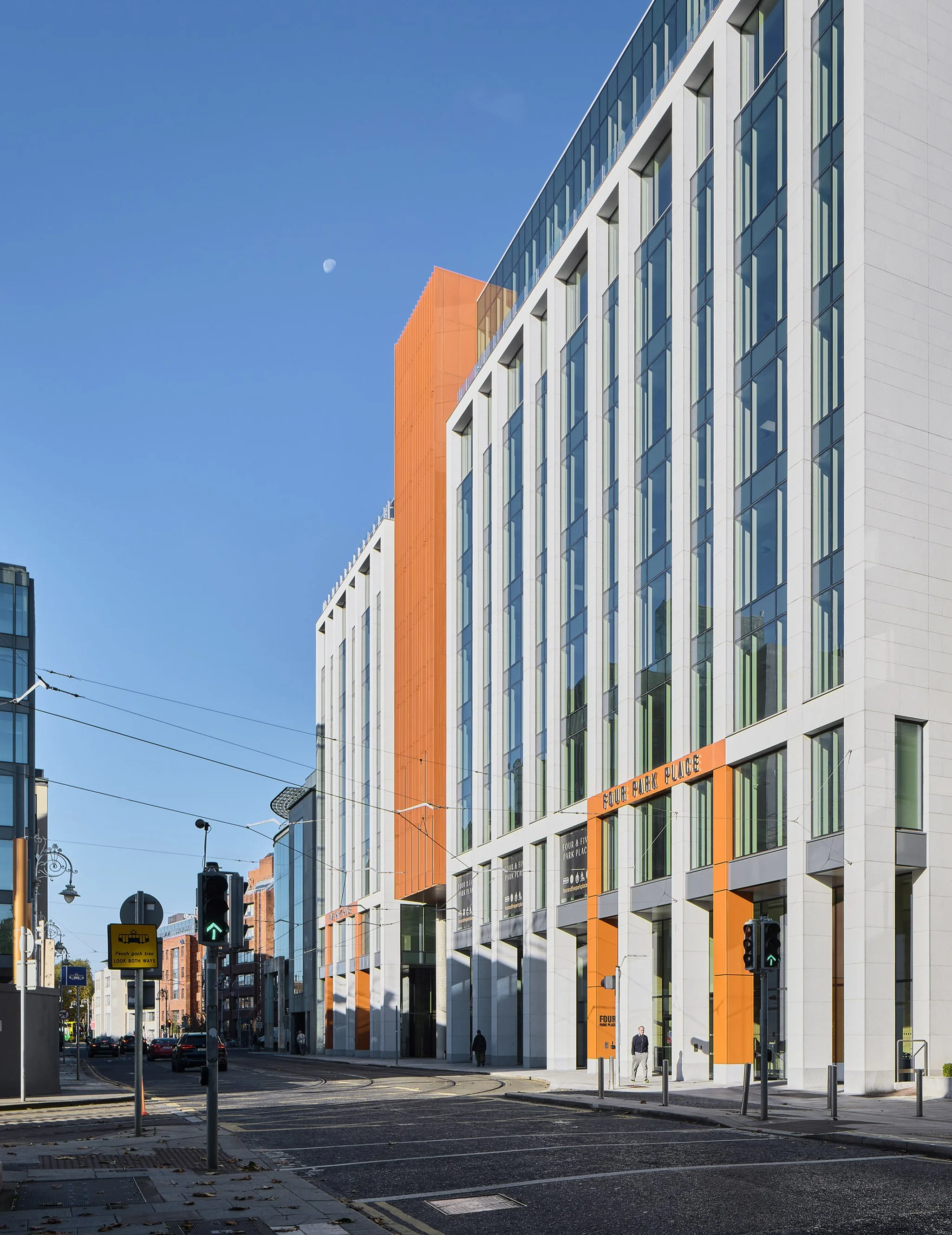Modern multi-story office building with white and orange exterior, large glass windows, and a street with traffic lights and pedestrians in front.