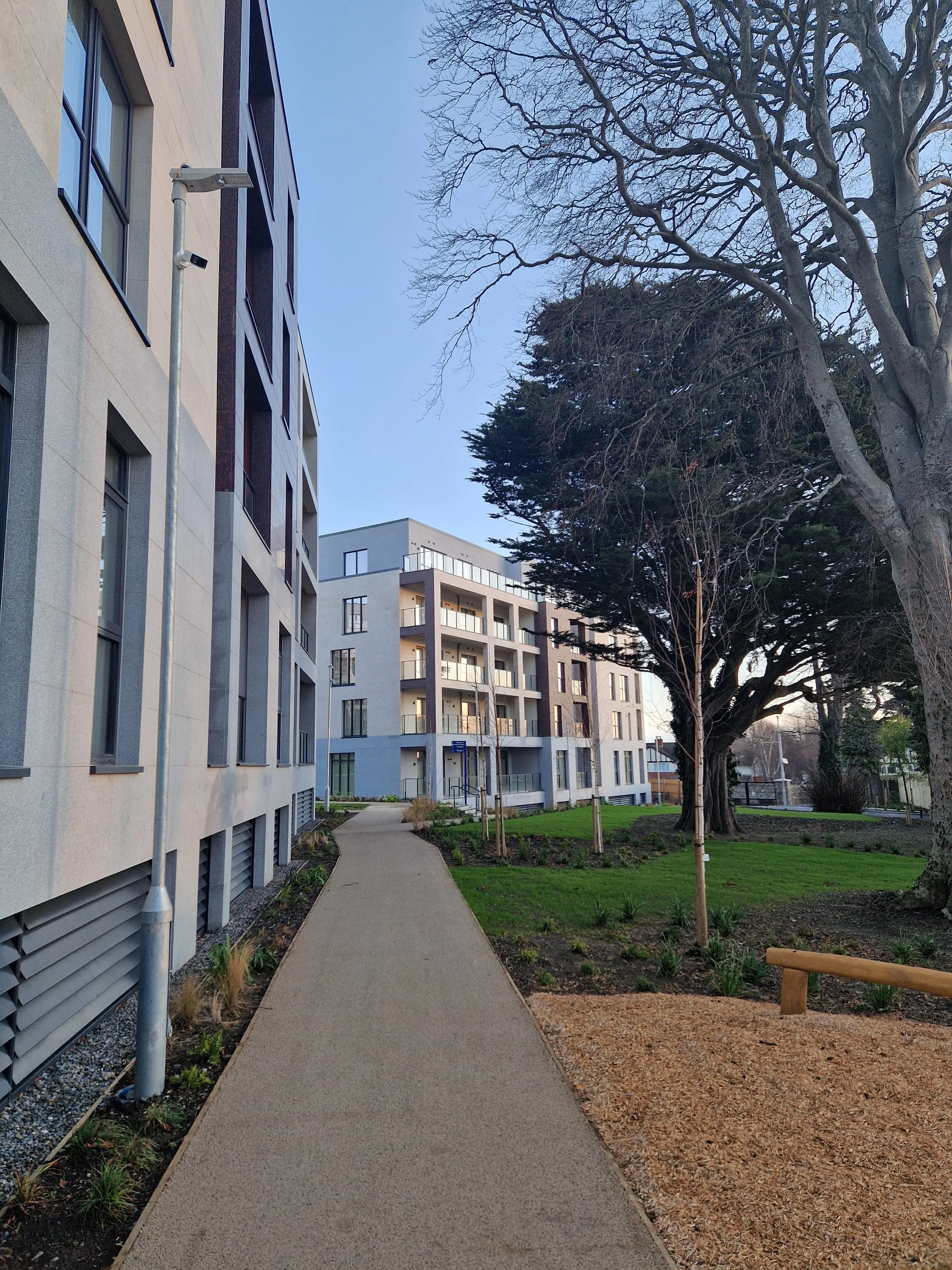 A pathway in a residential area with modern multi-story apartments on the left and trees on the right, during daytime with clear sky.