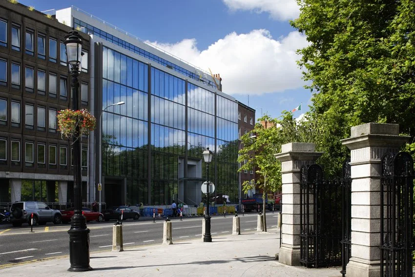 City street scene with modern glass building reflecting the sky, trees, and nearby buildings, black lamp posts, stone gate, parked cars, and sidewalk.