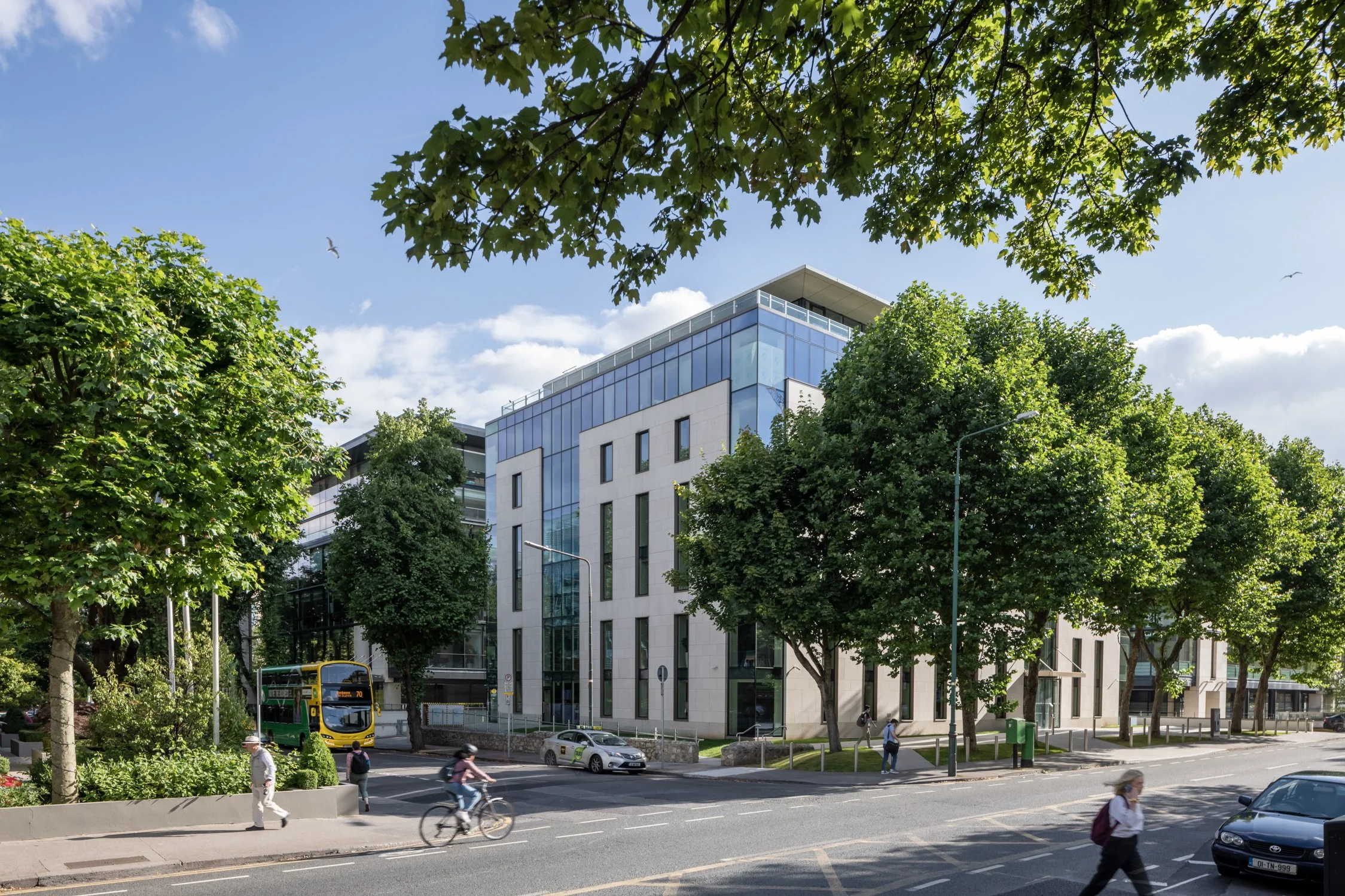 Modern office building with glass windows and white walls, surrounded by green trees, on a sunny day with some clouds, in an urban setting. Pedestrians are walking by, and a yellow double-decker bus and cars are on the street.