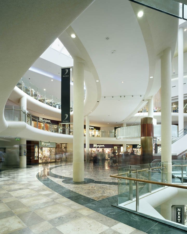 Interior of a modern shopping mall with multiple levels, glass railings, and several storefronts including Mangoon. There are blurred shoppers and a curved marble floor design.