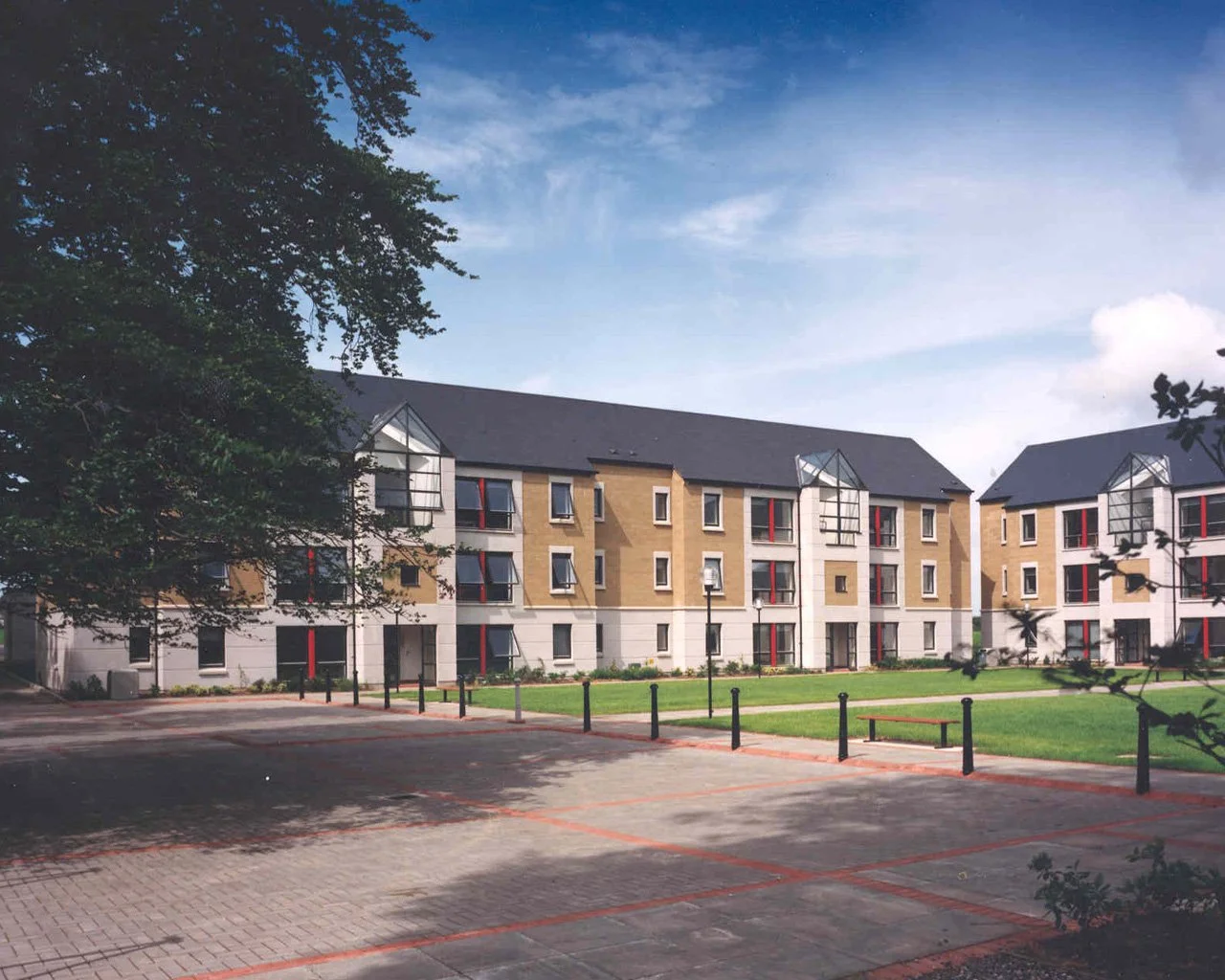 Modern multi-story apartment building with a parking lot and green lawn in front, trees on the sides, under a partly cloudy sky.