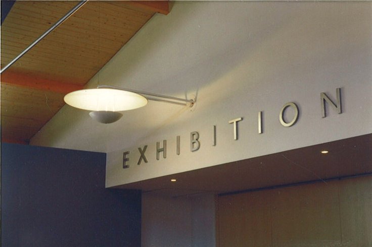 A sign reading 'EXHIBITION' in large metallic letters on a white wall above a dark-colored wall, in an indoor space with wooden ceiling panels and modern lighting.