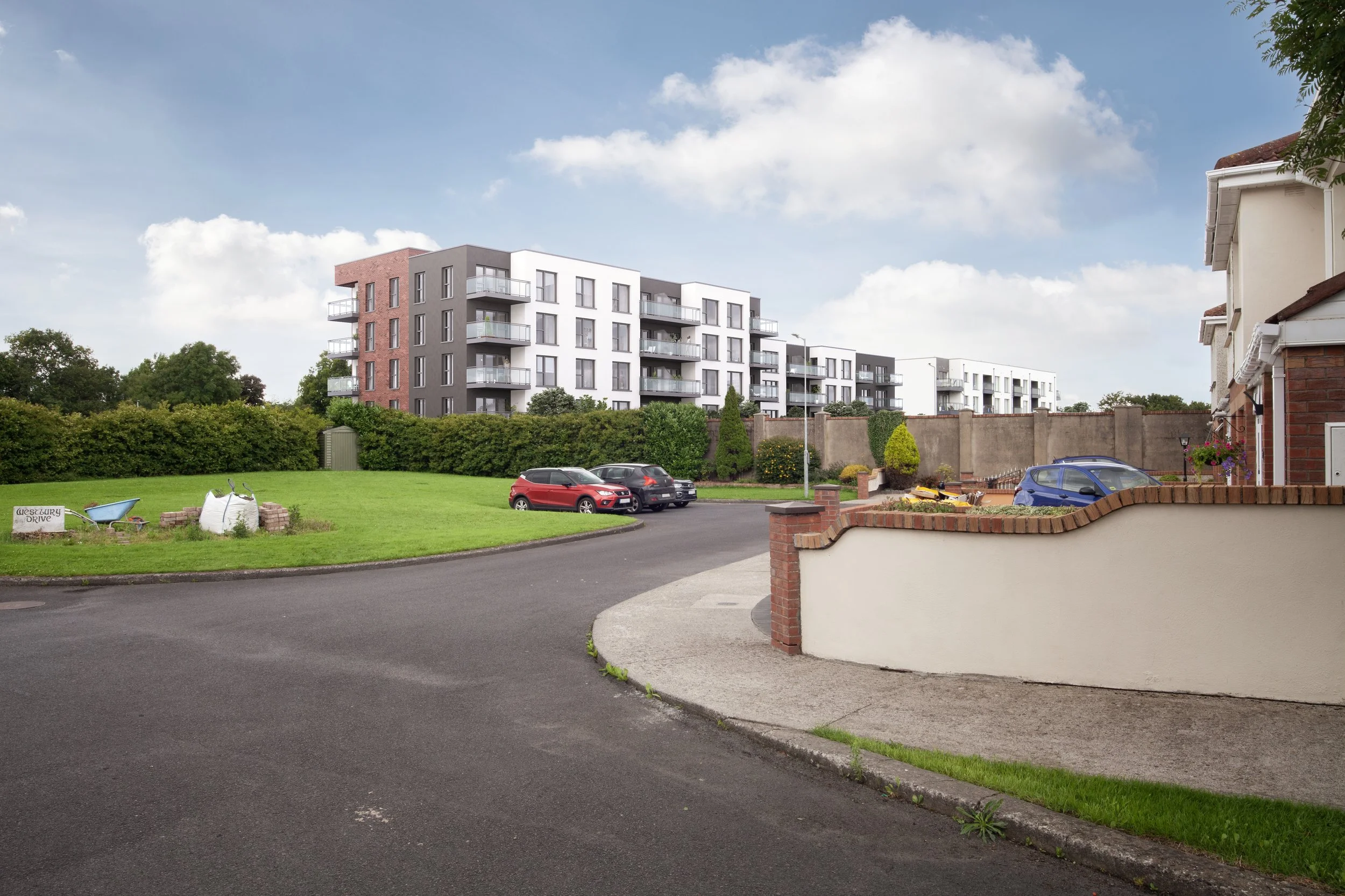 View of a suburban neighborhood with residential buildings, a small grassy area, and parked cars under a partly cloudy sky.