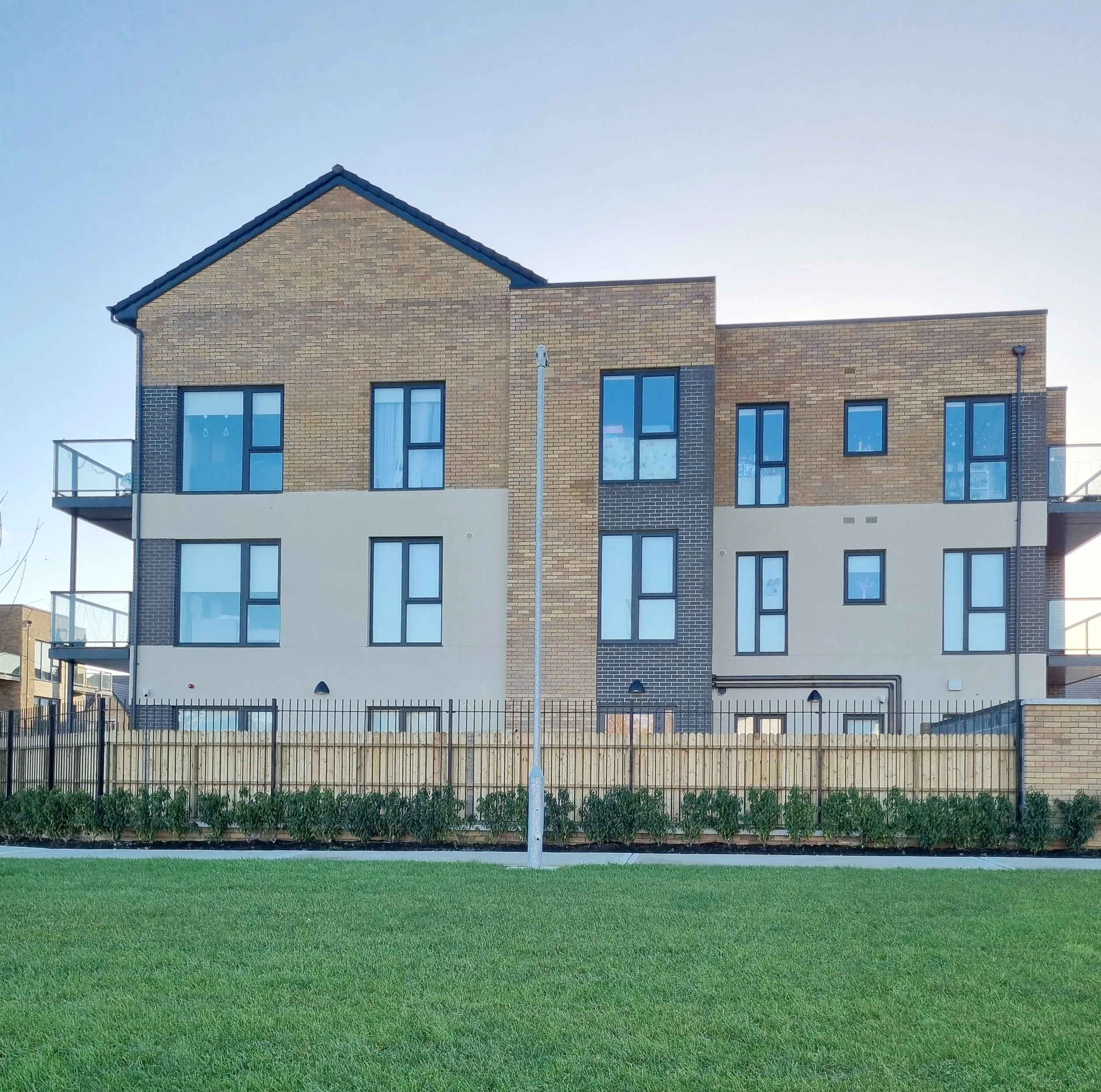 Modern multi-story apartment building with brick and beige exterior, multiple windows, and balconies, surrounded by a metal fence and green lawn in the foreground.
