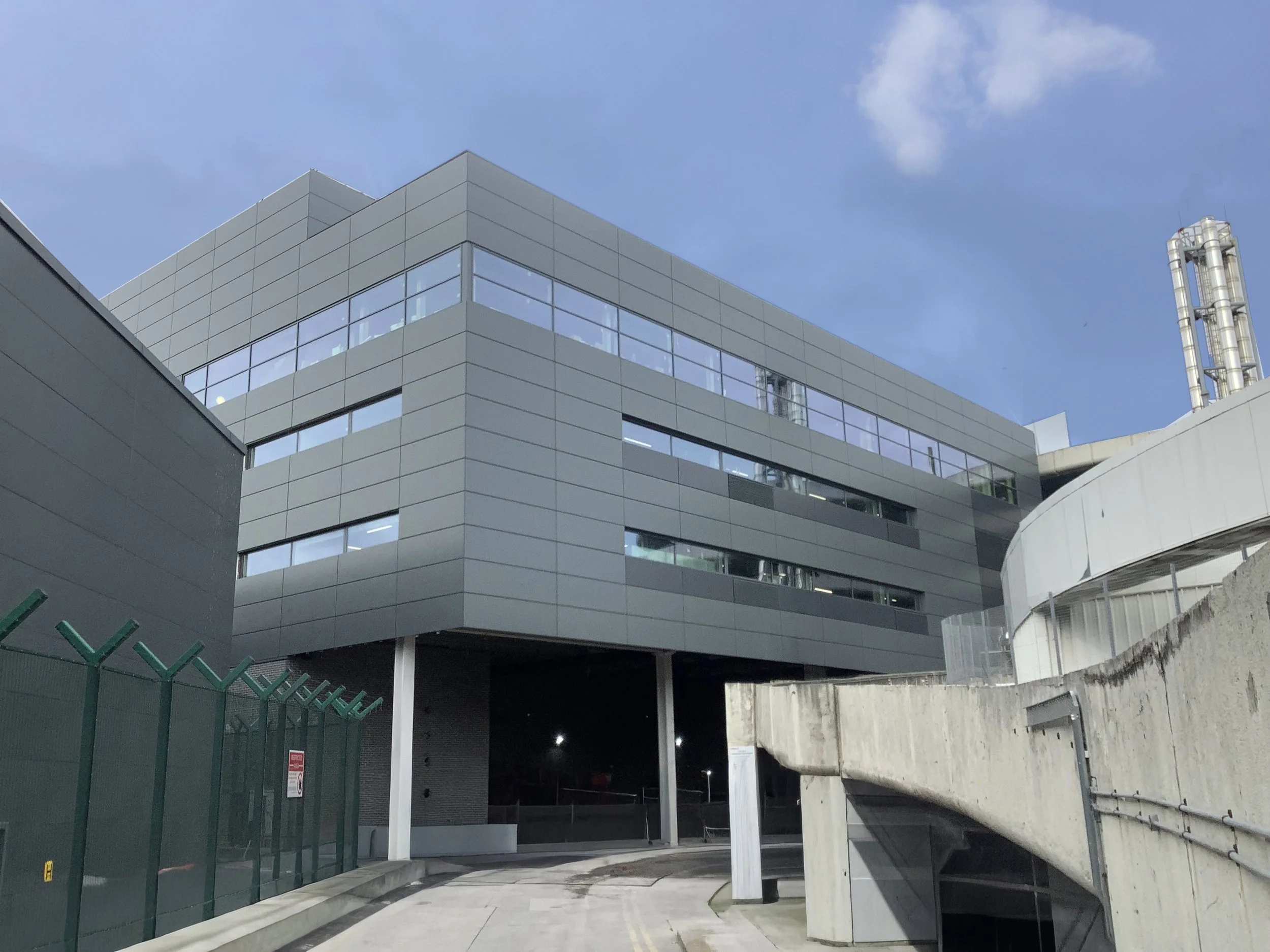 Modern gray building with large windows and surrounding concrete structure, under a partly cloudy sky.
