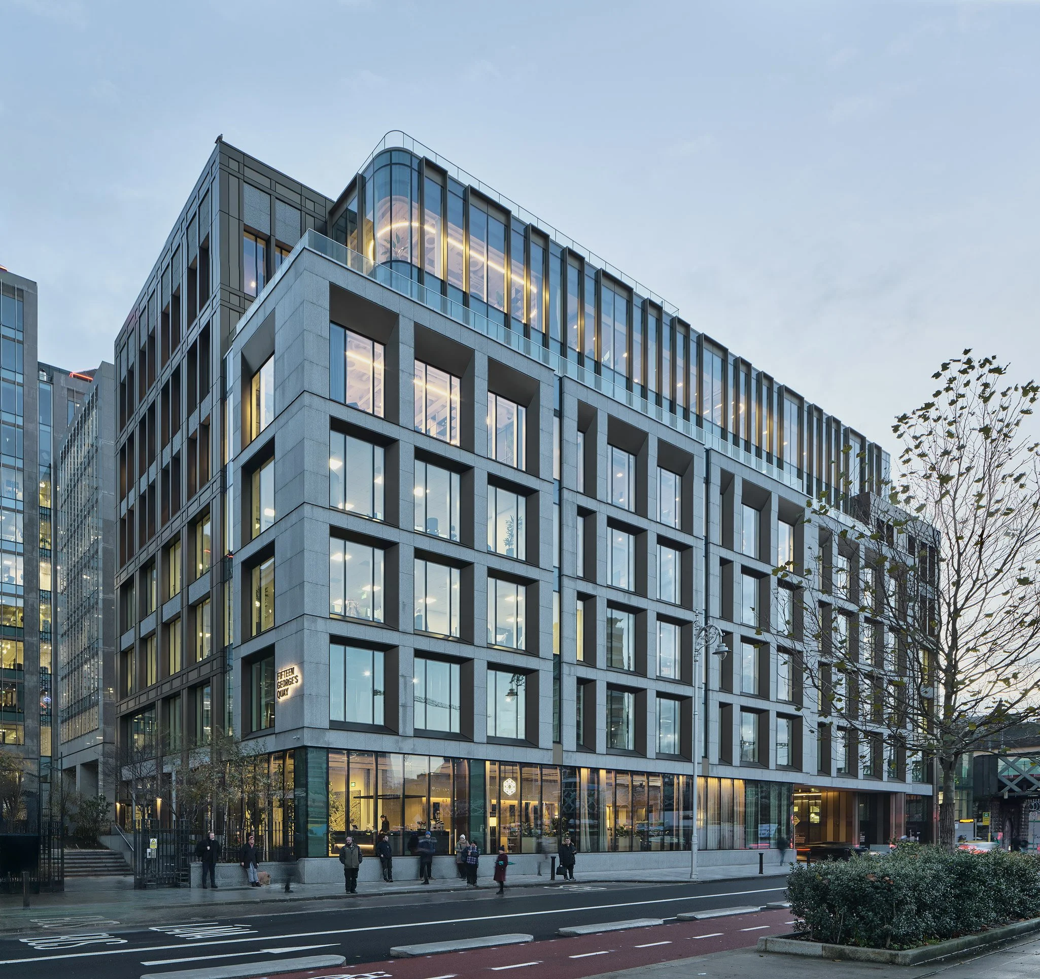 Modern multi-story office building with large glass windows and a ground-floor lobby, with people walking on the sidewalk in front, and a tree on the right side, during evening or twilight.