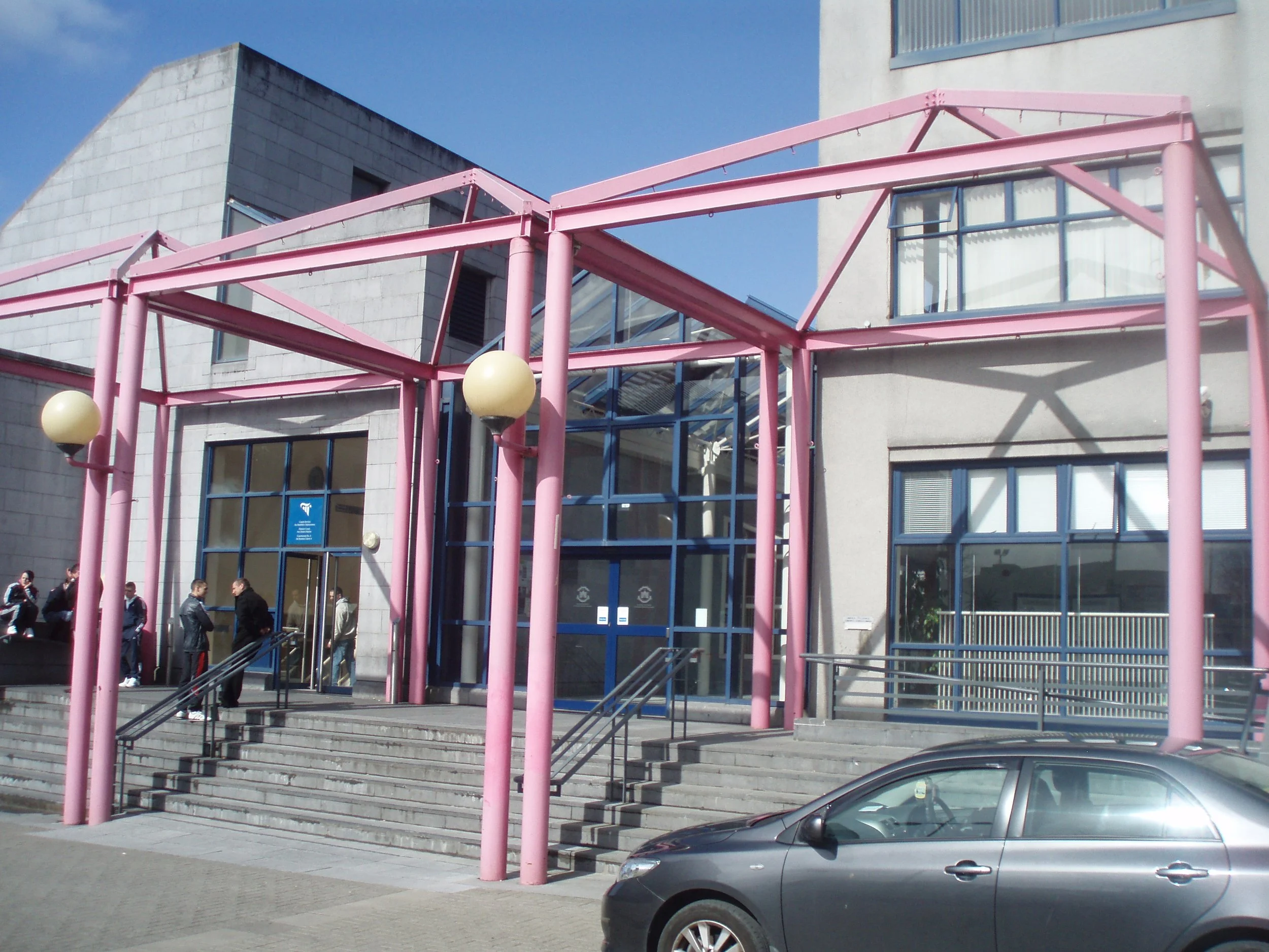 Exterior of a modern building with large glass windows and pink metal framework at the entrance, with several people standing near the entrance and a parked gray car in the foreground.