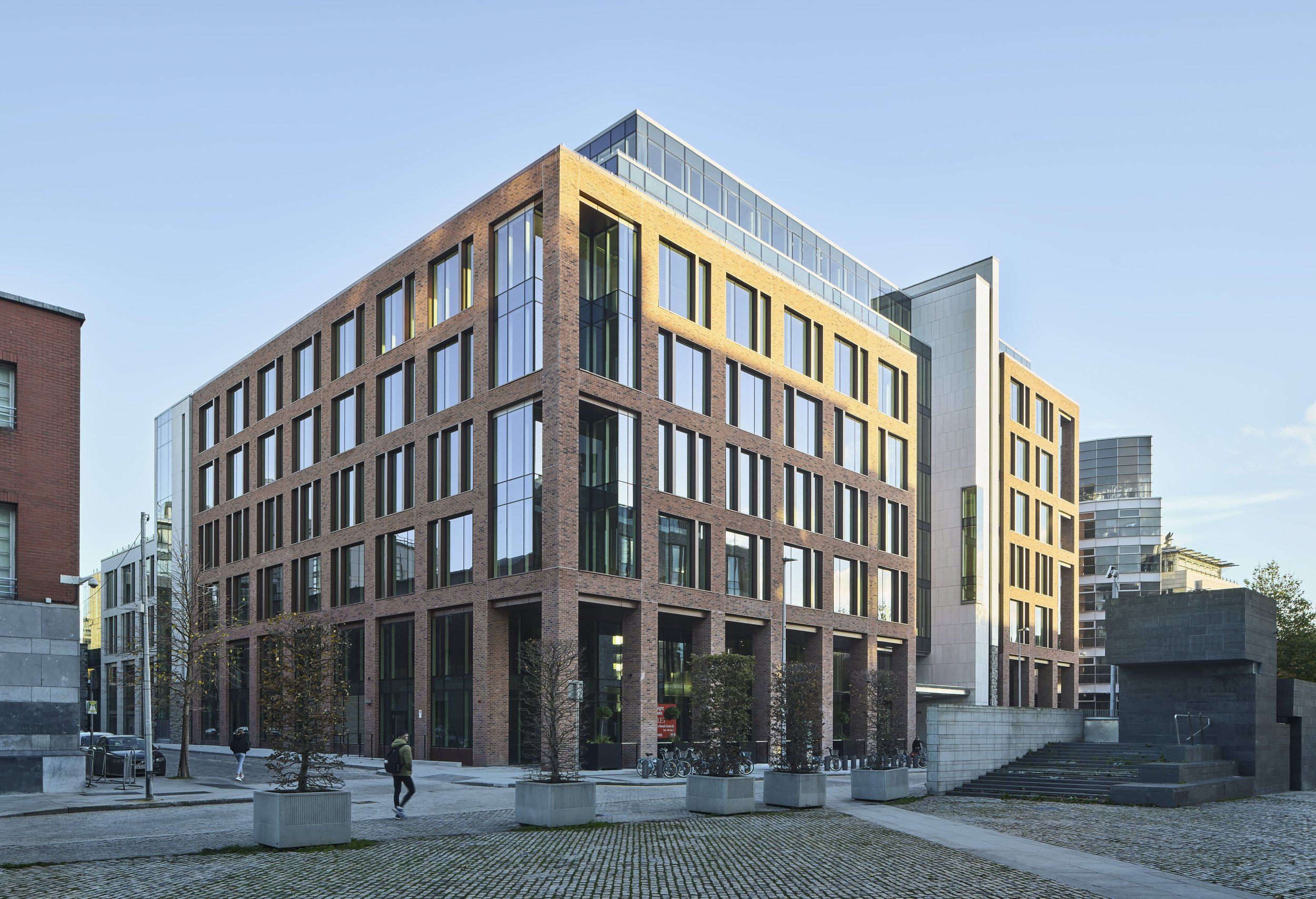 Modern multi-story office building with brick and glass facade, situated in an urban area with pedestrians and potted trees.