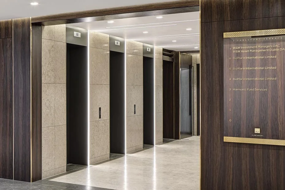 Four modern elevator doors in a sleek building lobby with wood and stone interior design and an directory sign on the wall.