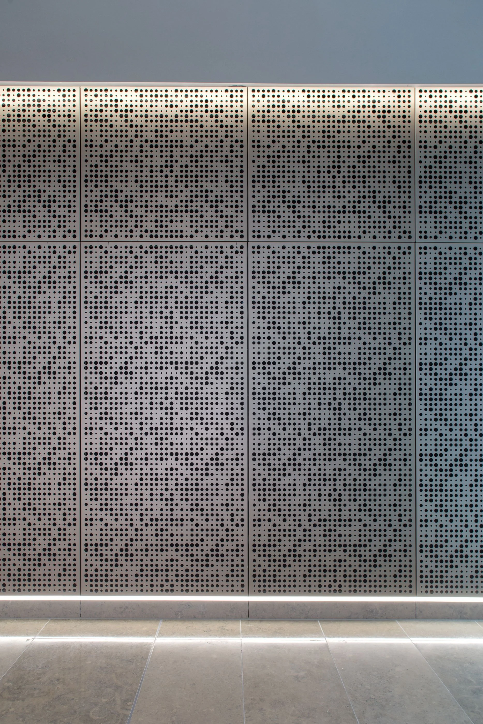 Close-up of a gray perforated metal wall with evenly spaced round holes, illuminated by a linear light at the bottom, with a tiled floor.